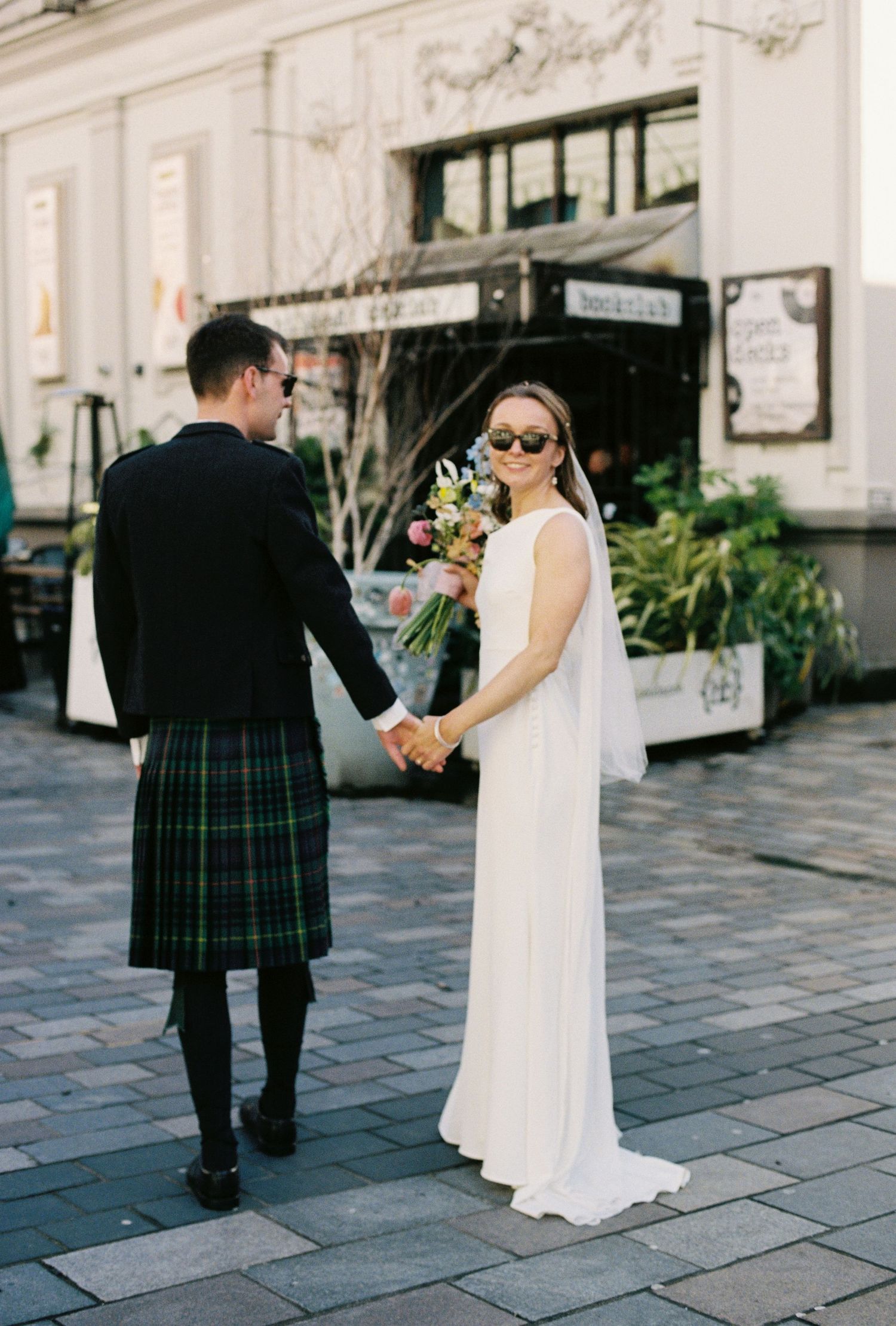 Bride in white gown and groom in tartan kilt hold hands on cobblestone street, bride holding colorful bouquet.
