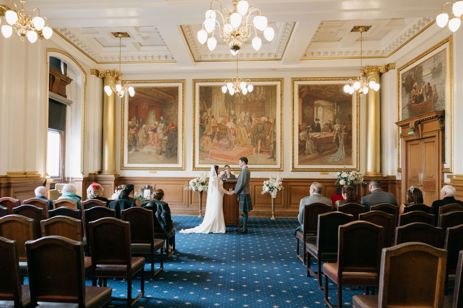 Intimate wedding ceremony in an ornate Edinburgh City Chambers room with blue carpet and historic paintings.