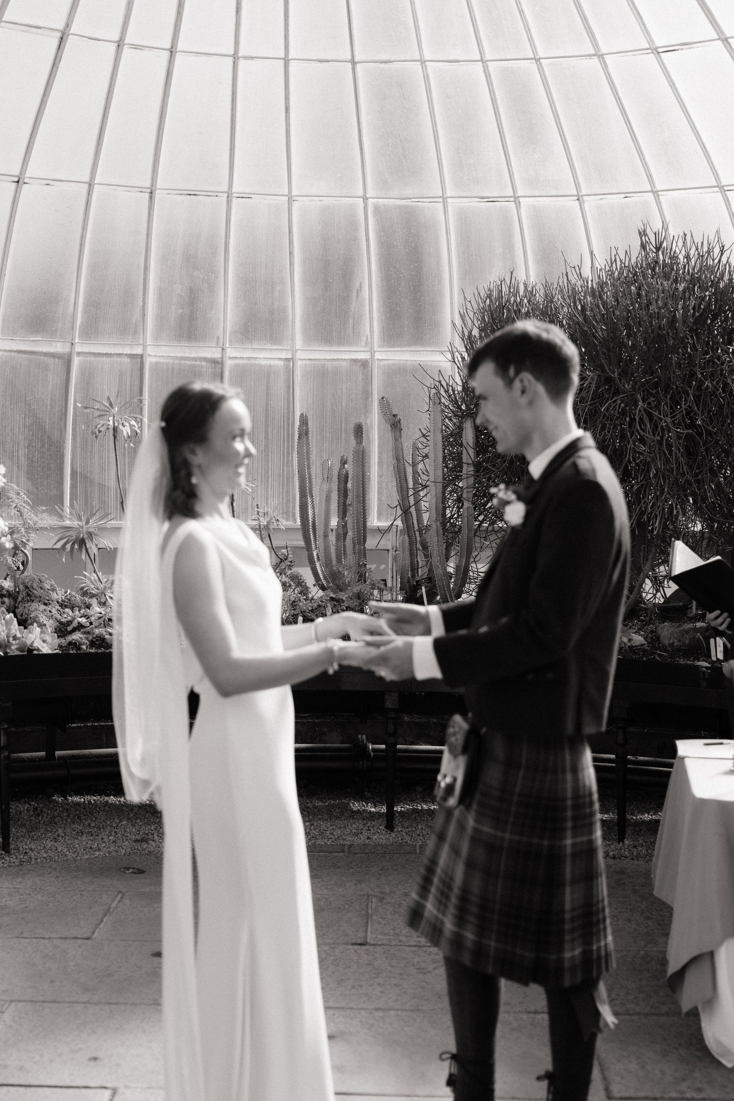 Black and white photo of a bride and groom exchanging vows in a stunning glass greenhouse wedding ceremony.