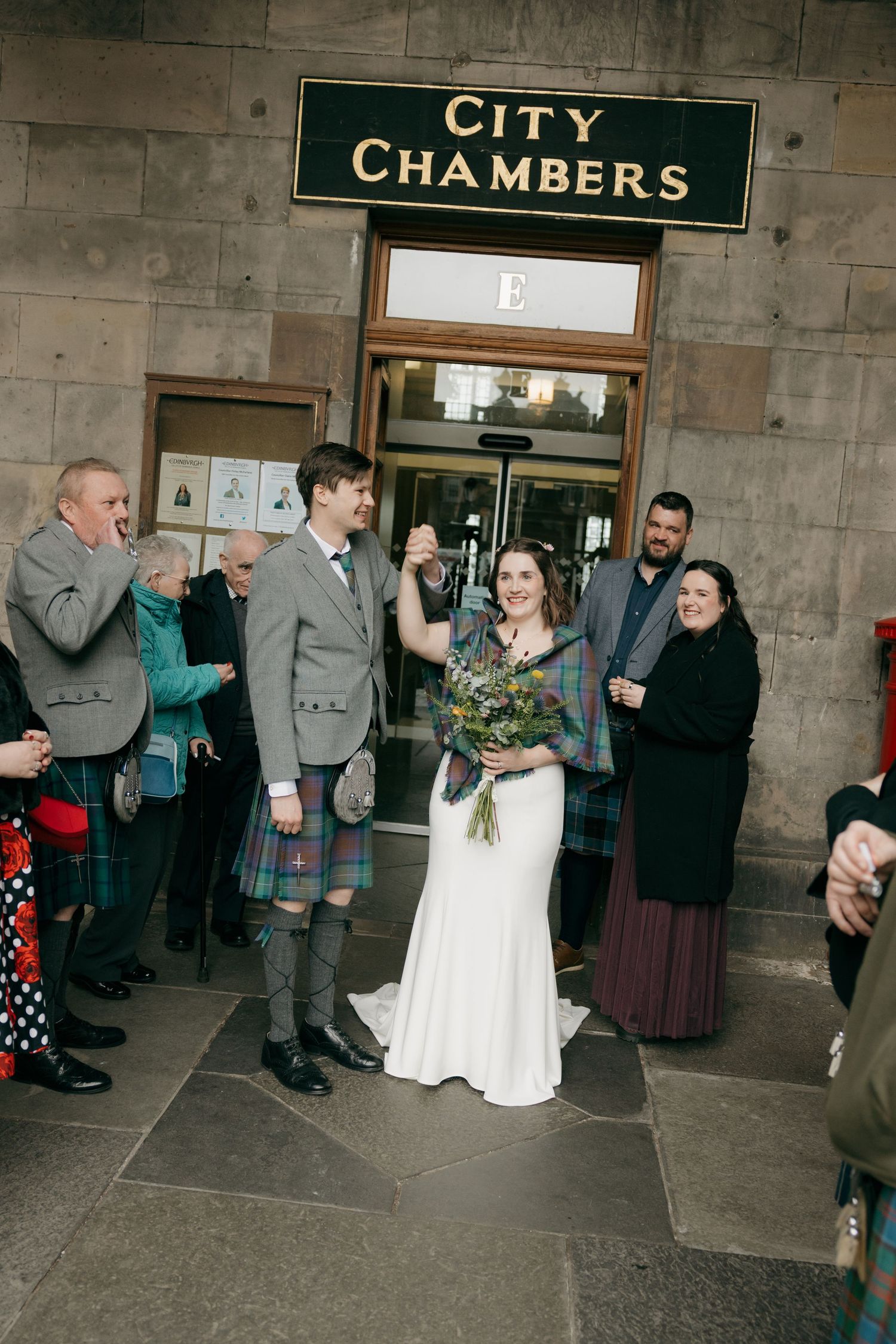 Newlyweds celebrate outside Edinburgh City Chambers, groom in tartan kilt, bride holding bouquet.