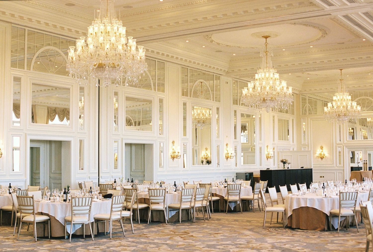 Elegant grand ballroom with crystal chandeliers, white chiavari chairs, and round dining tables set for a formal event.