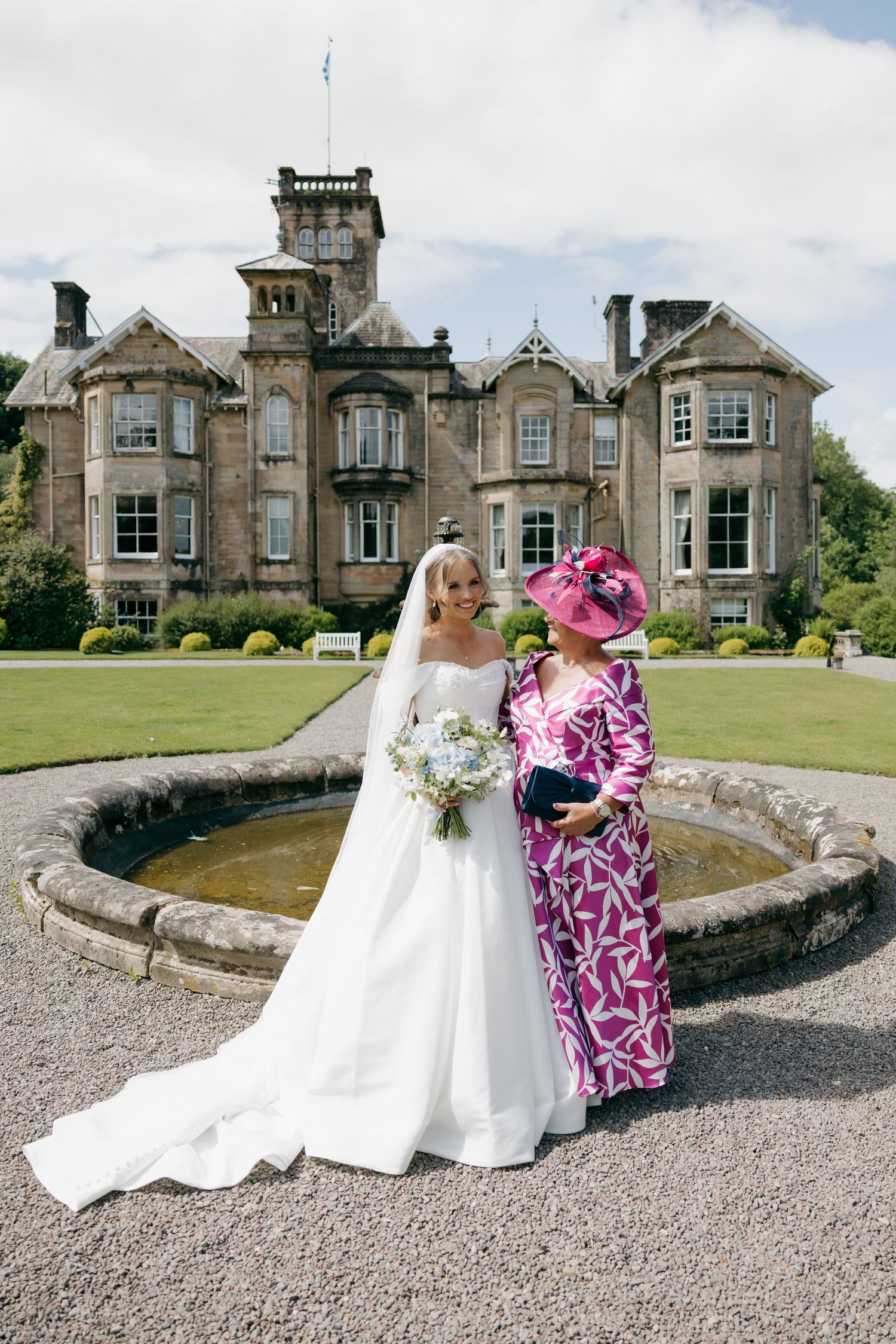Bride in white gown with bouquet and guest in pink hat pose by fountain outside Scottish castle venue.