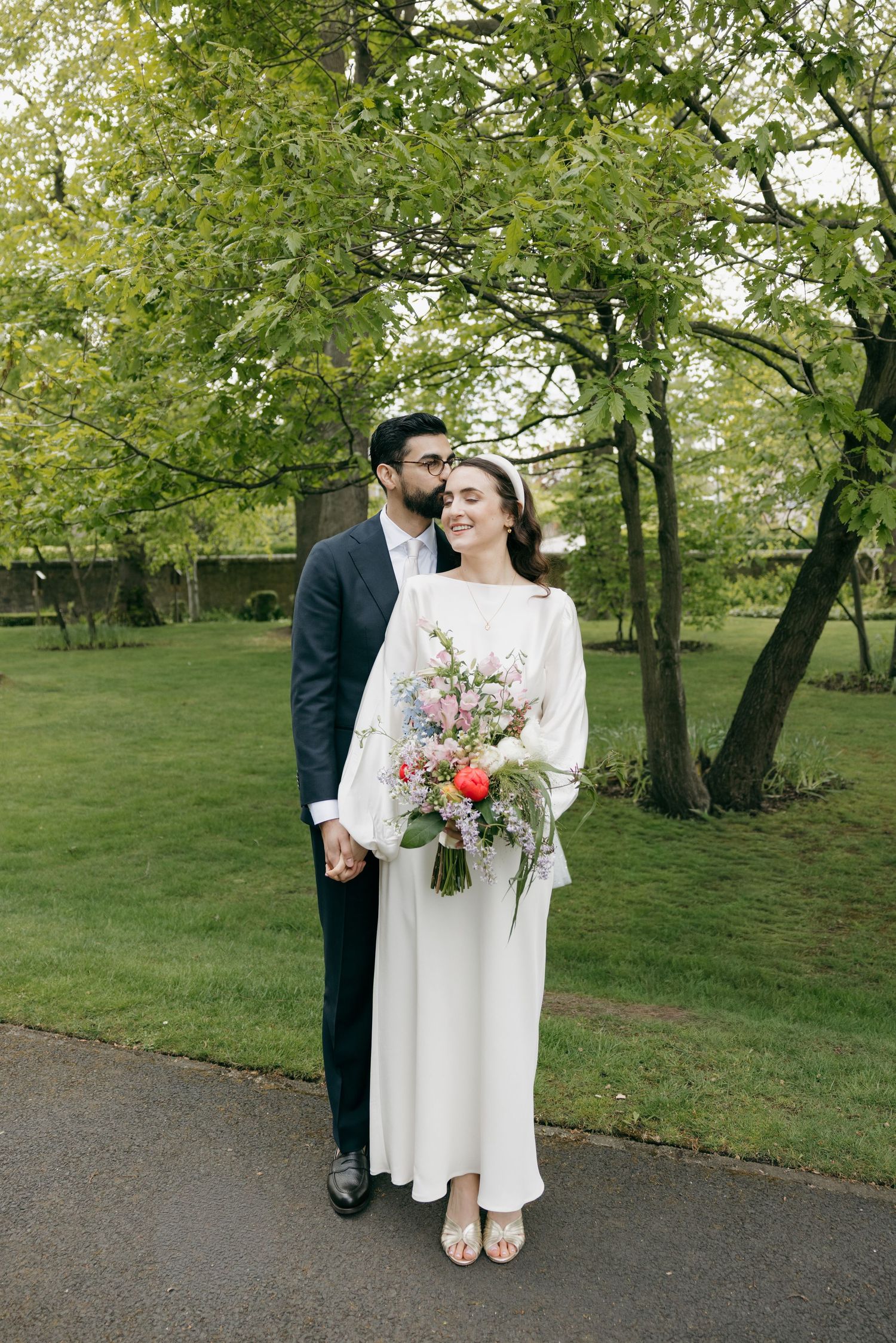 A bride and groom hold hands in a lush green park, she holds a colorful bouquet in a white gown, he wears a dark suit.