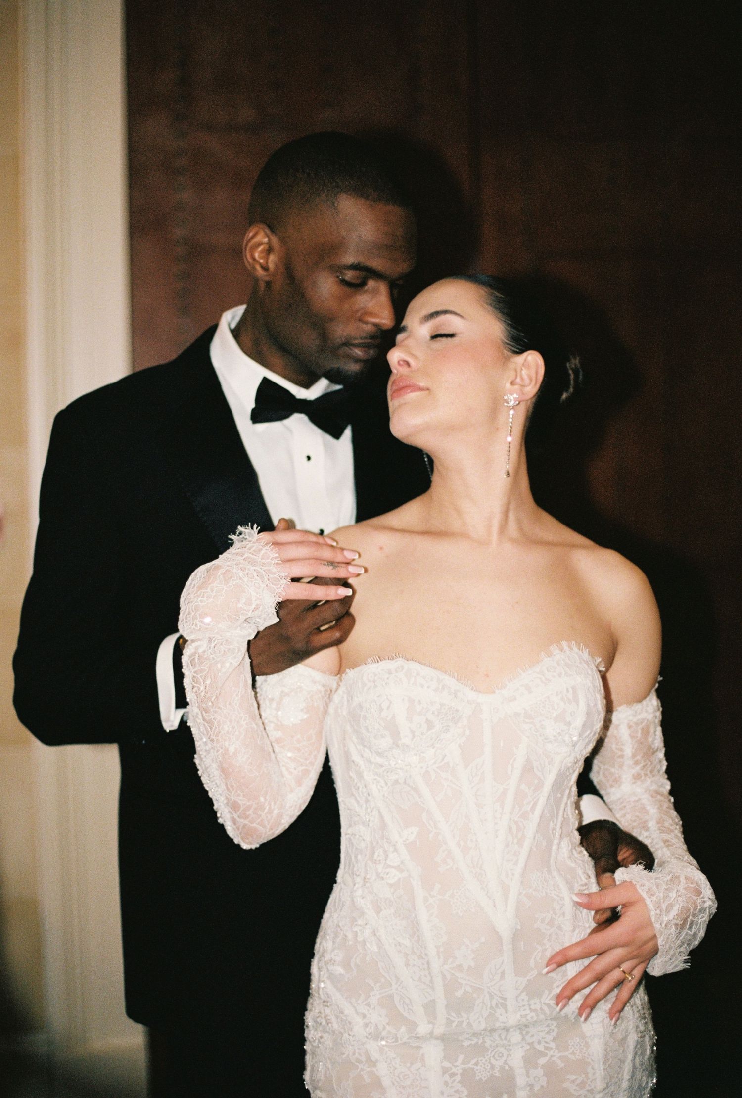 A well-dressed couple embracing in a formal setting, with the man in a tuxedo and the woman in a white wedding gown.