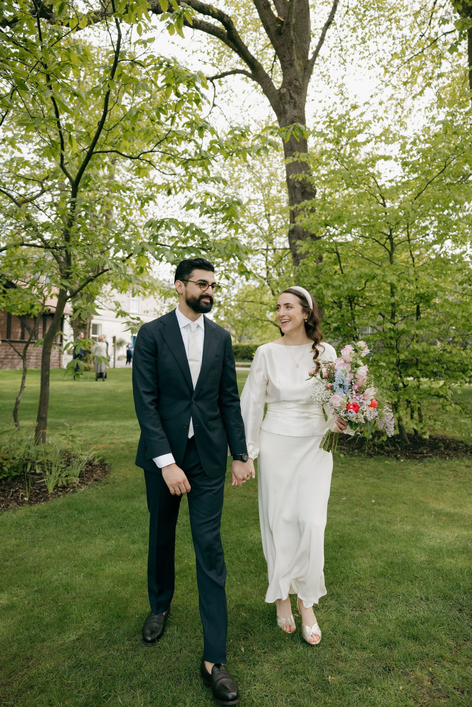A couple in wedding attire holds hands while walking through a lush green park, bride holding a colorful bouquet.