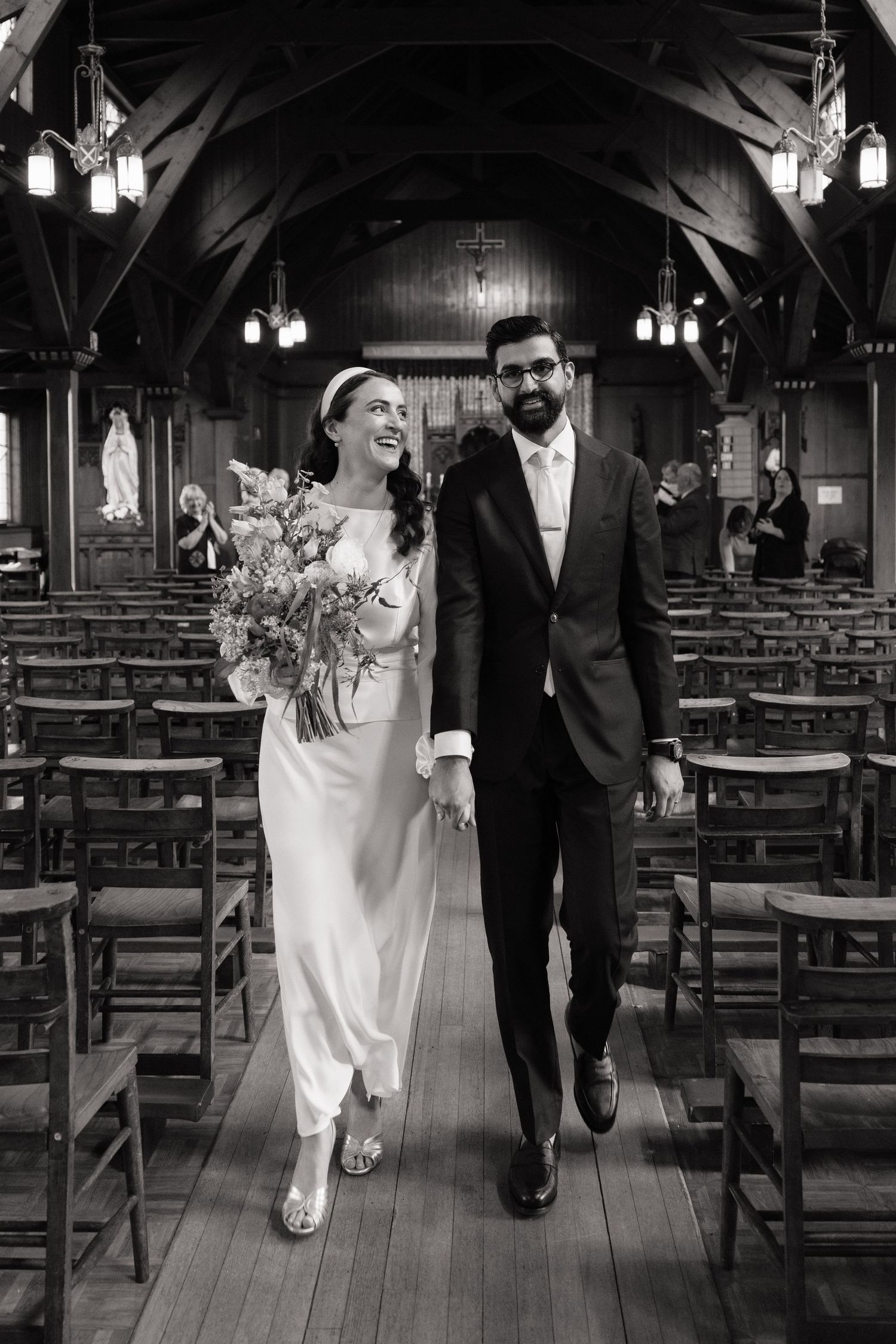 Black and white photo of a smiling bride and groom walking down the aisle of a wooden church after their wedding ceremony.