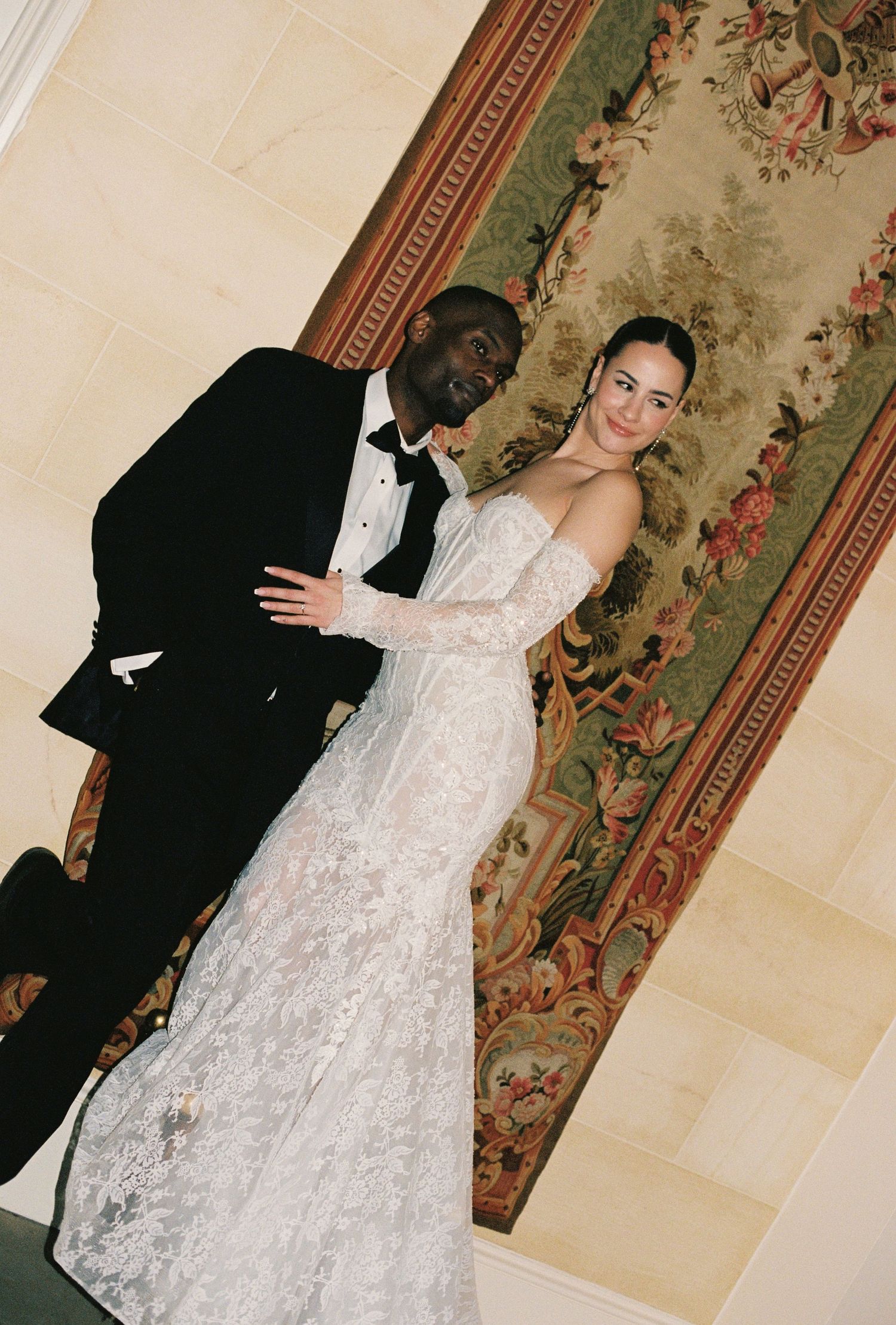 A man in a black tuxedo and a woman in a white lace wedding dress pose together in a formal setting.