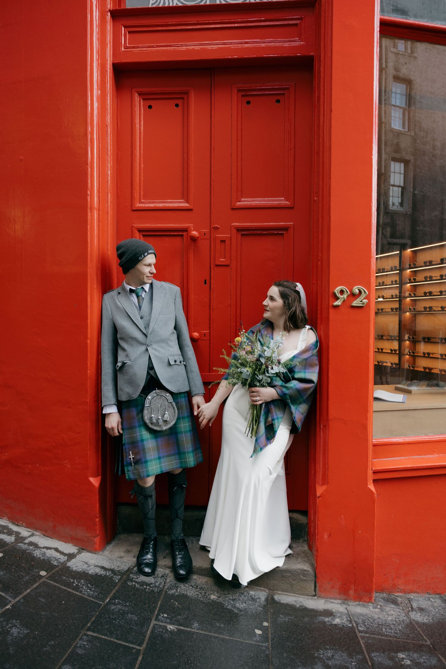 Couple in Scottish wedding attire holding hands before a red door in Edinburgh, groom in tartan kilt, bride in white gown.