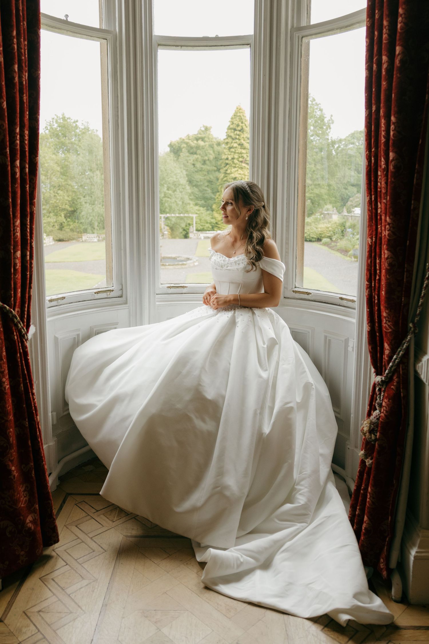 Bride in off-shoulder ballgown standing in bay window of elegant venue with garden views.