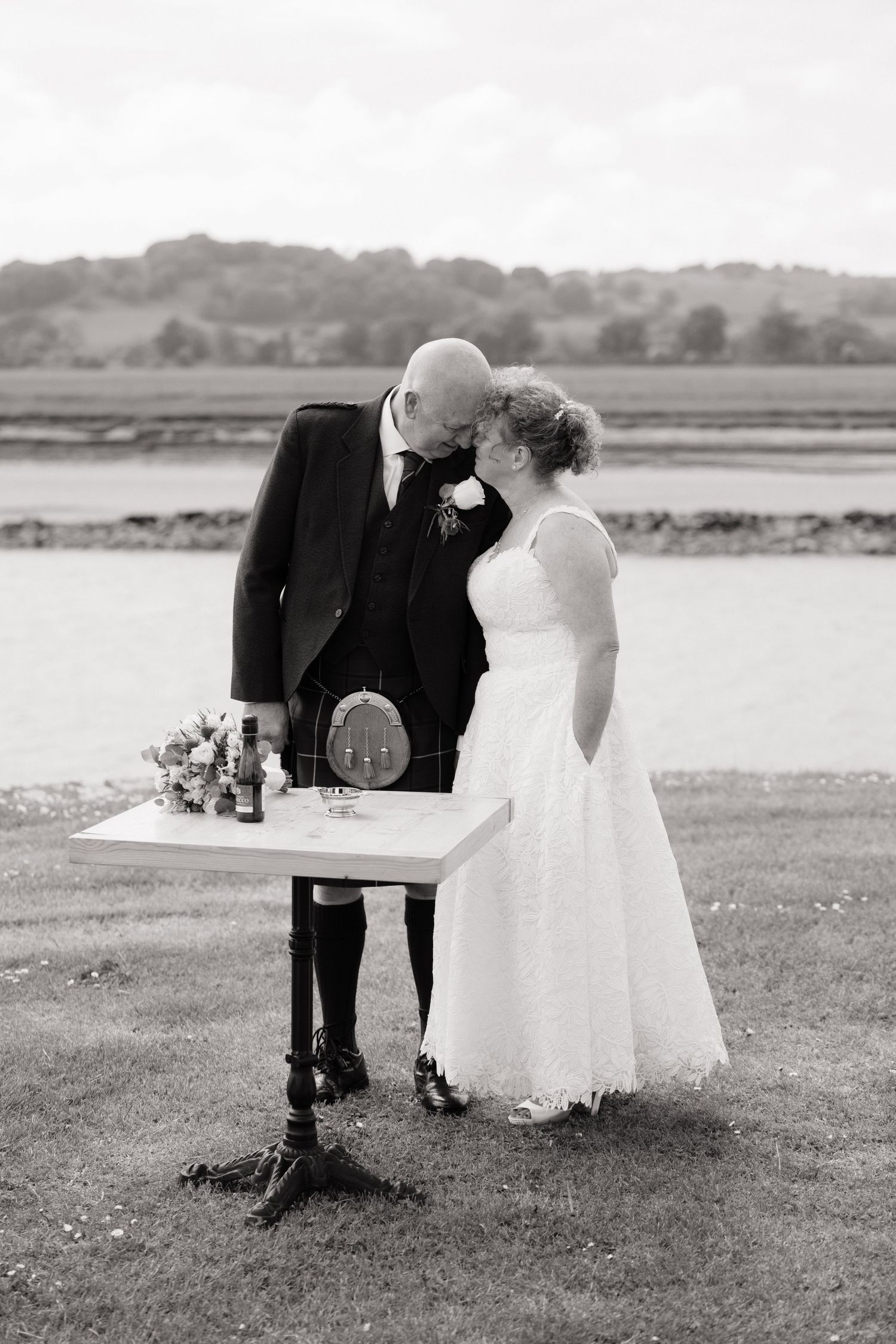 Black and white photo of a bride and groom sharing a tender moment at their outdoor wedding ceremony by a waterfront.