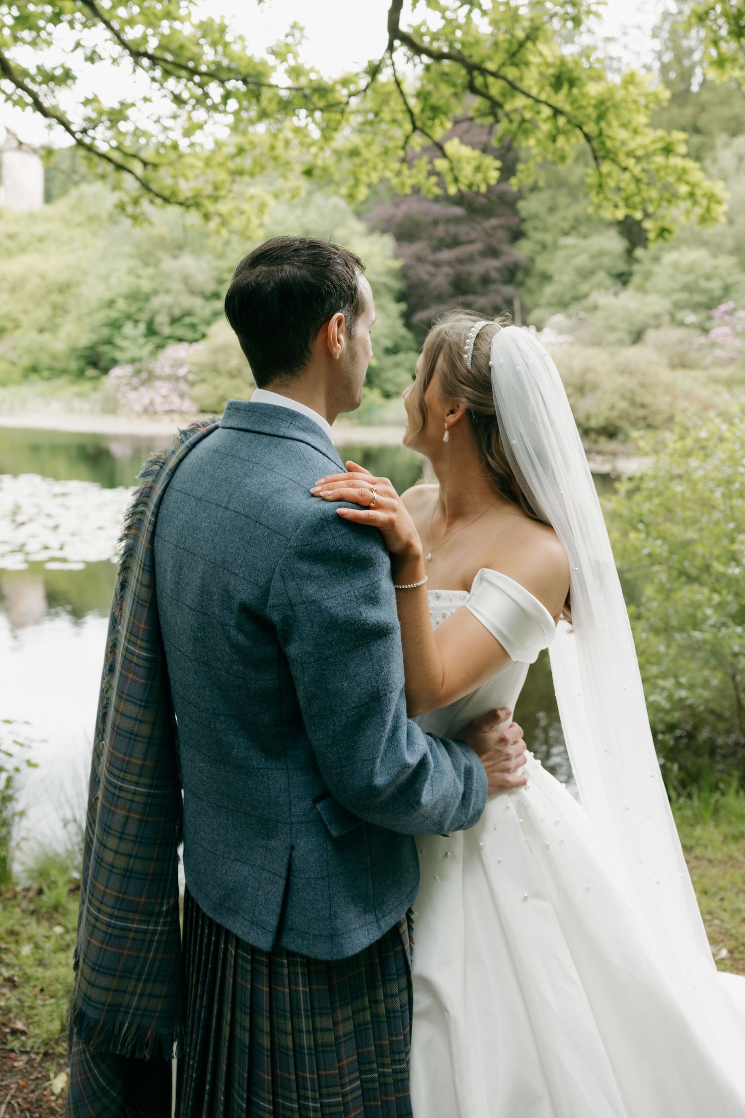 Bride and groom embrace facing a serene garden lake, bride in off-shoulder gown with long veil.