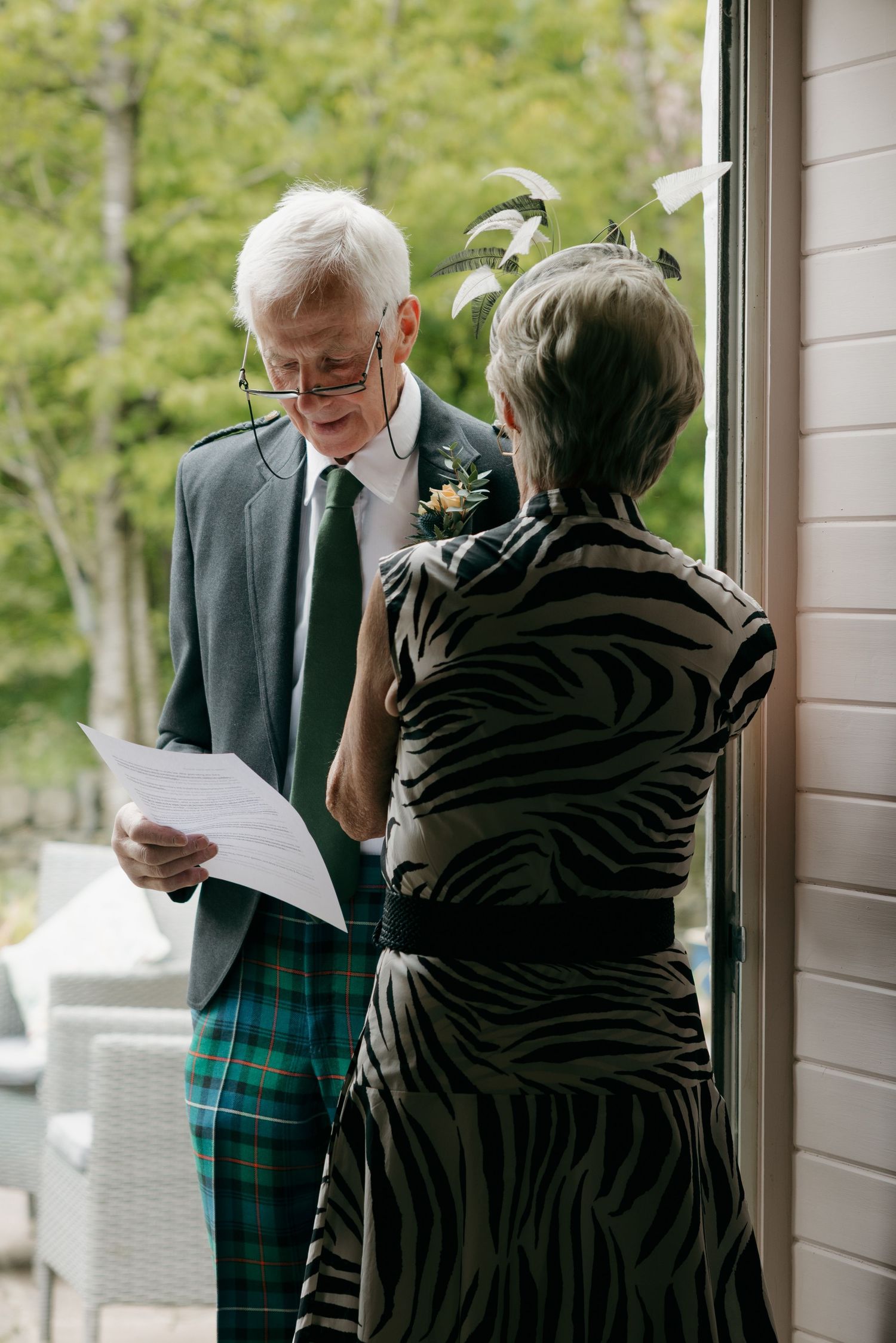 Elderly couple in formal attire reviewing wedding ceremony papers, man wearing tartan kilt and boutonniere.