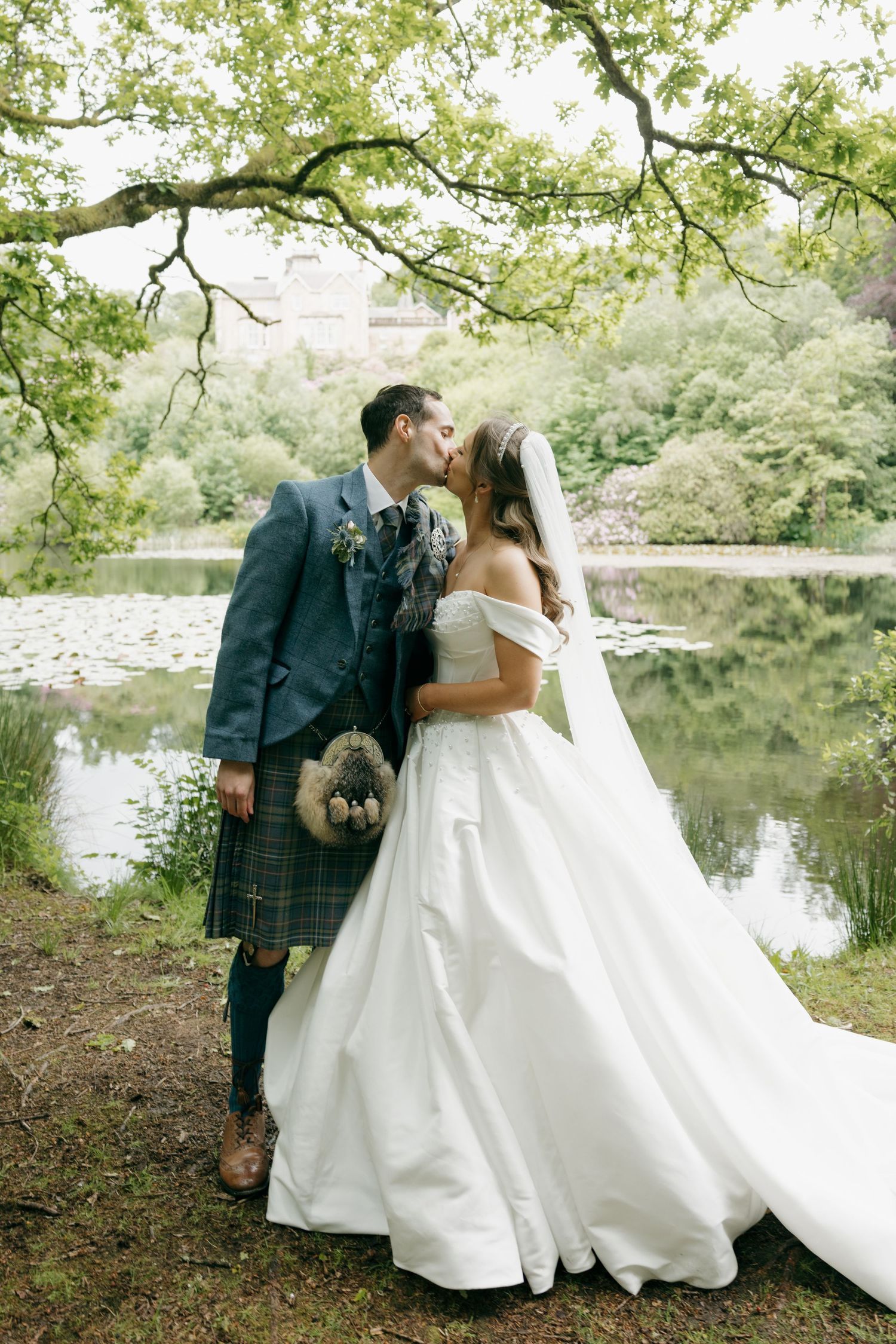 Newlyweds share romantic kiss beside peaceful lake surrounded by lush greenery on wedding day.