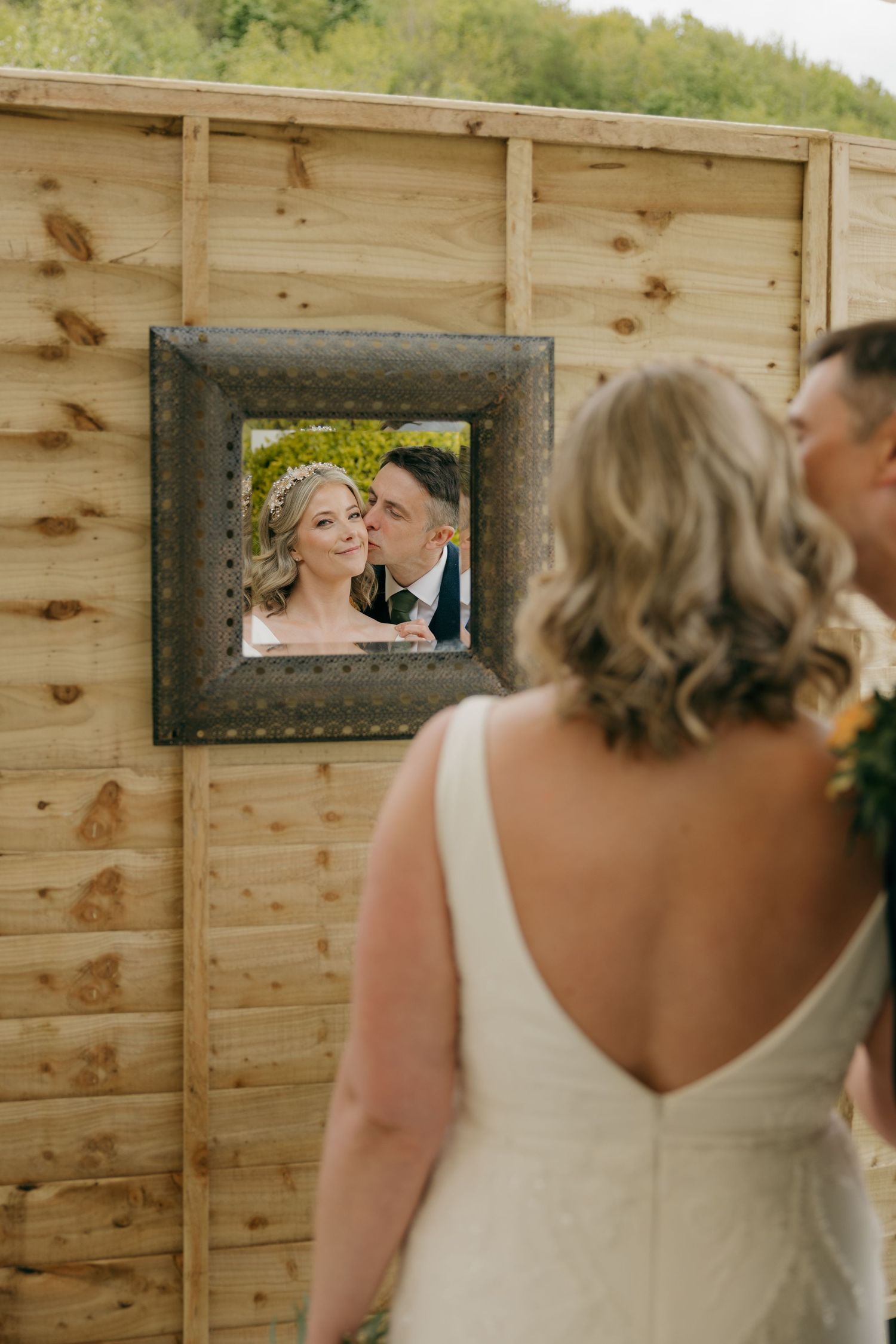 Bride in white gown views framed wedding photo of couple kissing on rustic wooden wall backdrop.