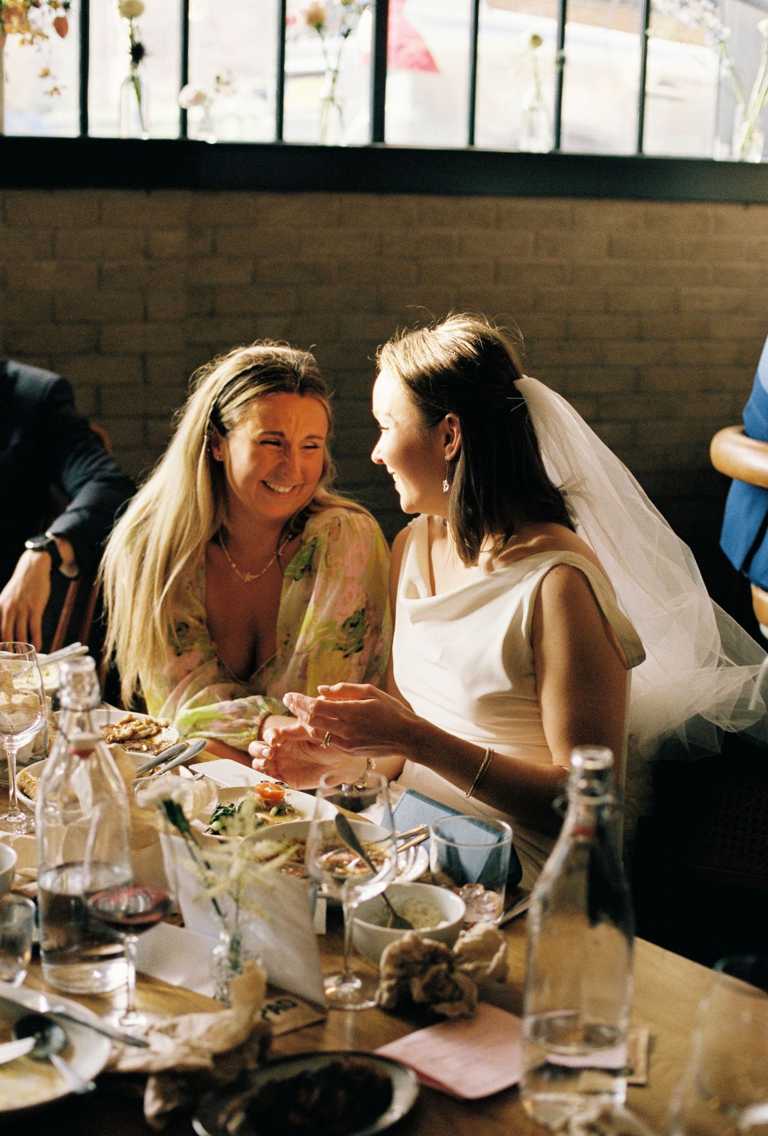 A bride in a white dress and veil shares a joyful moment with a blonde guest at a wedding reception dinner.