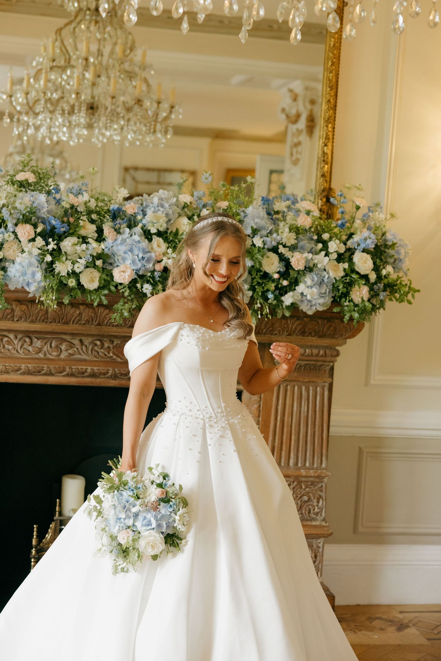 Smiling bride in off-shoulder white ball gown holding blue bouquet, standing before ornate gold fireplace with floral arrangements.