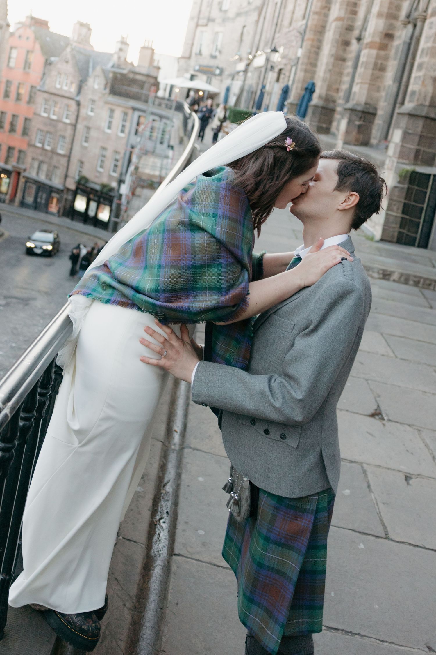 A bride and groom share a romantic kiss on Edinburgh's Royal Mile, he wearing a traditional Scottish kilt and she in a white gown.