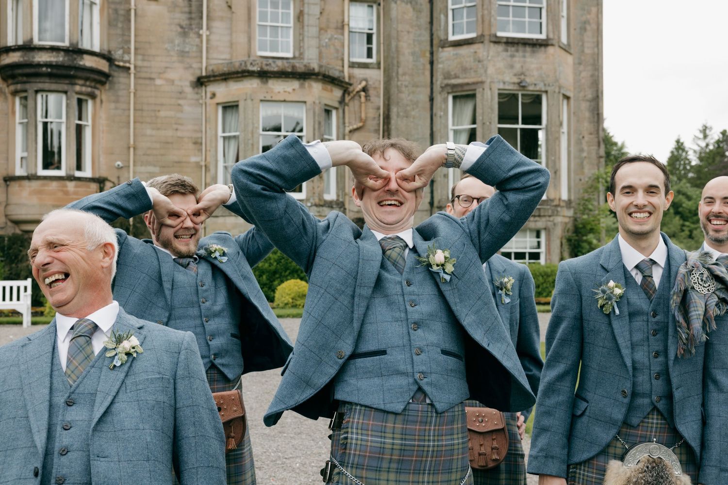 Groomsmen in blue suits and tartan kilts laughing and posing outside a Scottish country house wedding venue.