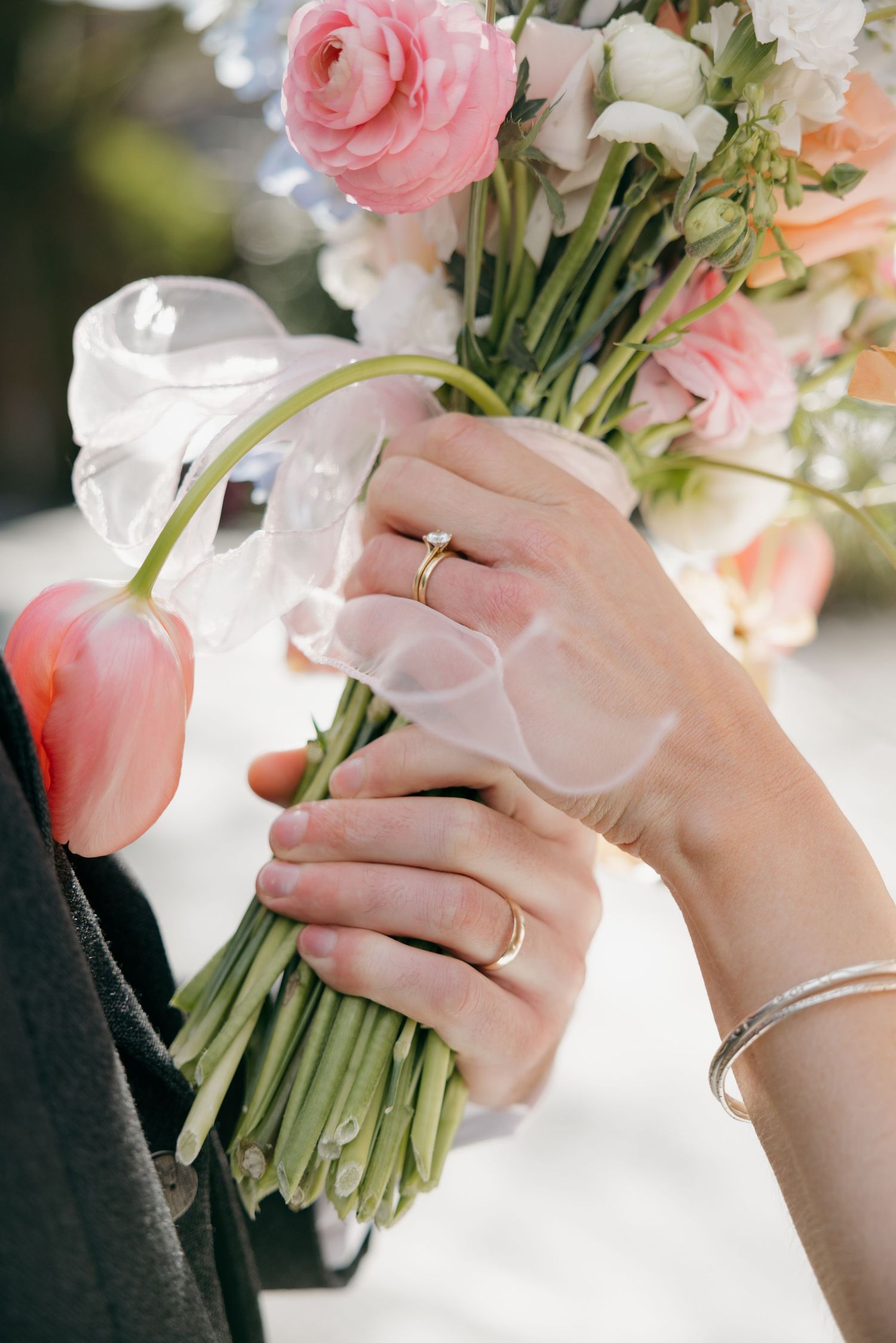 Bride and groom holding a pastel wedding bouquet with pink tulips and ranunculus, bride wearing gold diamond ring and silver bracelet.