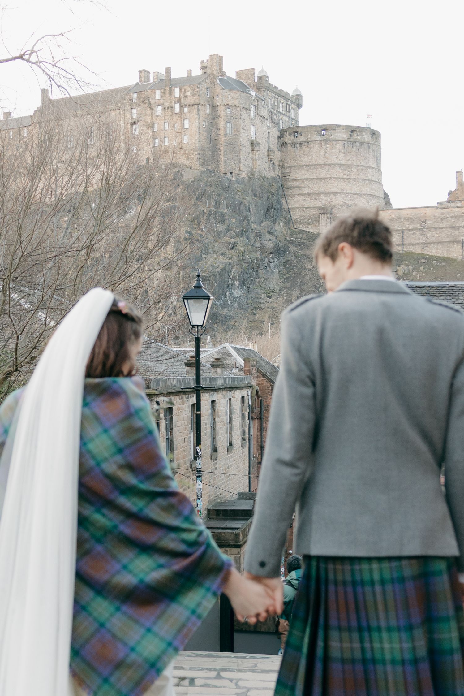 A bride and groom in tartan attire hold hands while gazing at Edinburgh Castle on their wedding day.