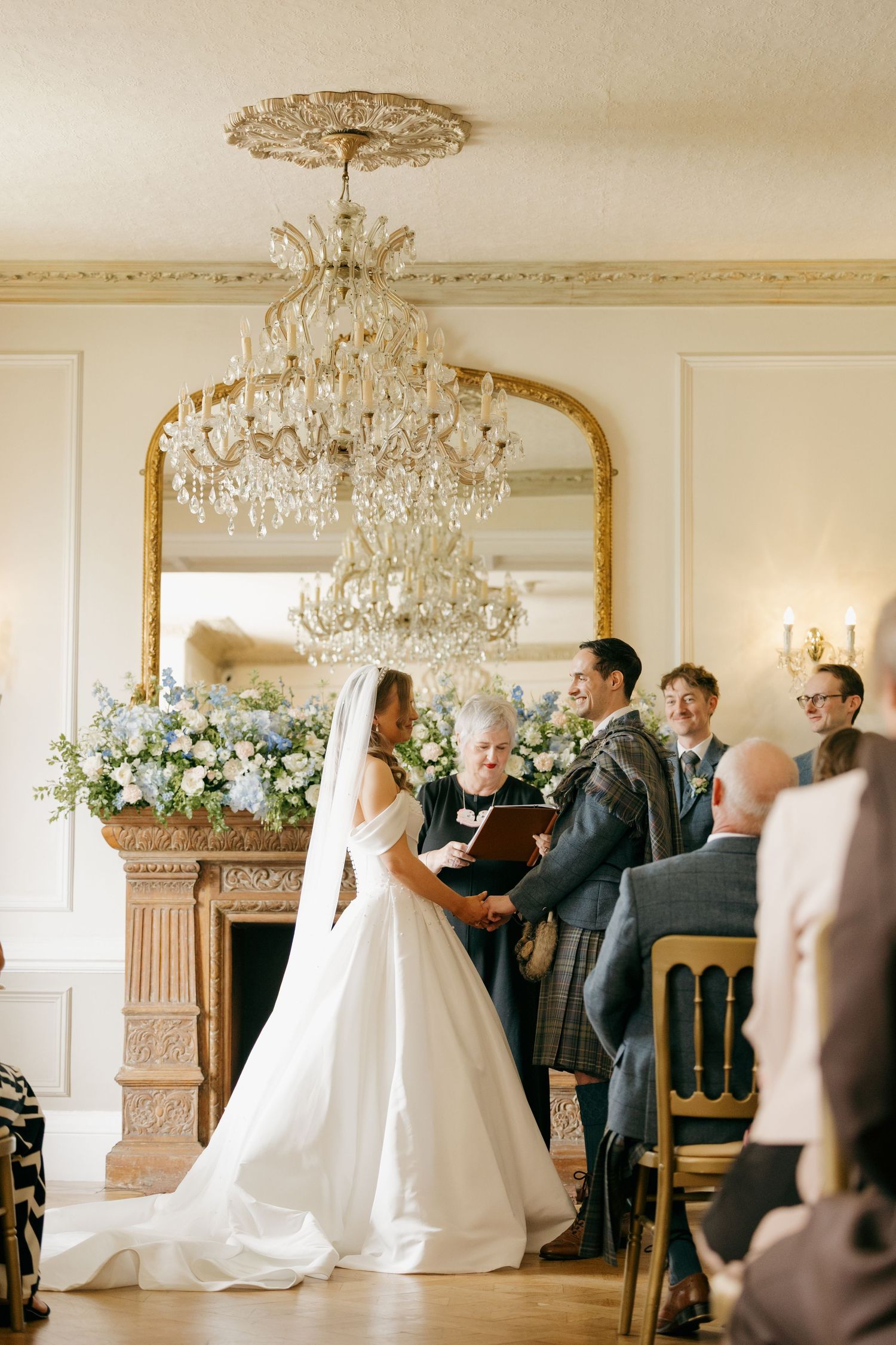 Bride and groom exchange vows beneath crystal chandelier with gold mirror and blue floral arrangements.