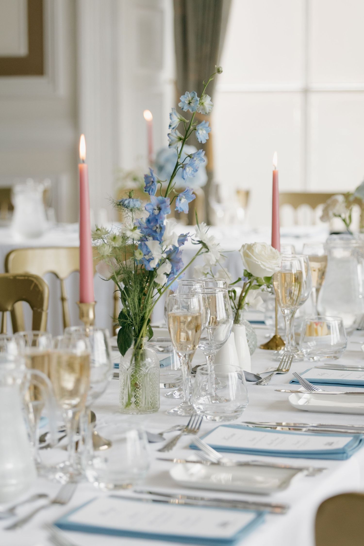 Elegant wedding reception table set with blue flowers, pink candles, champagne glasses, and white linens.