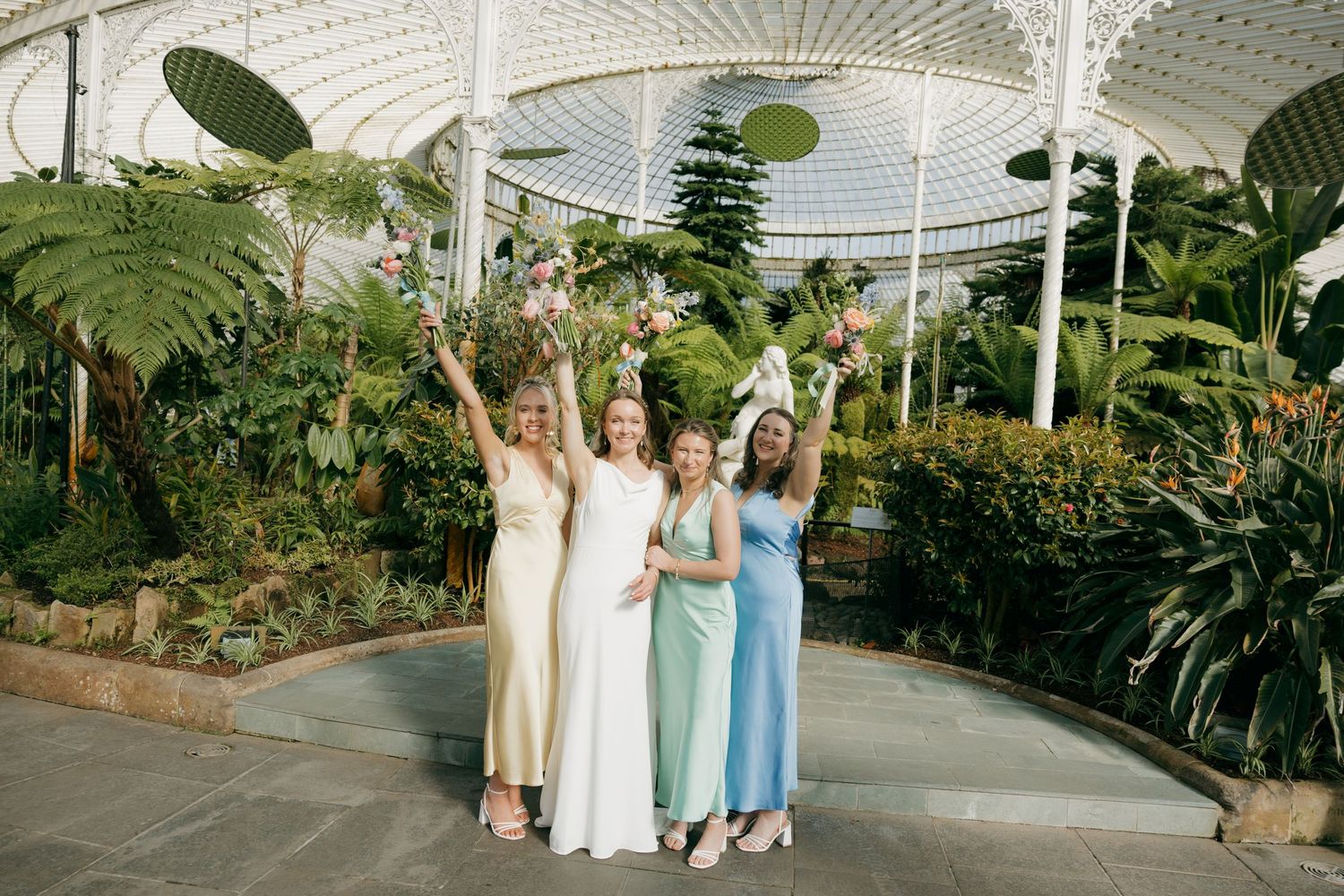 Bride and bridesmaids in colorful dresses celebrating inside a lush botanical garden greenhouse venue.