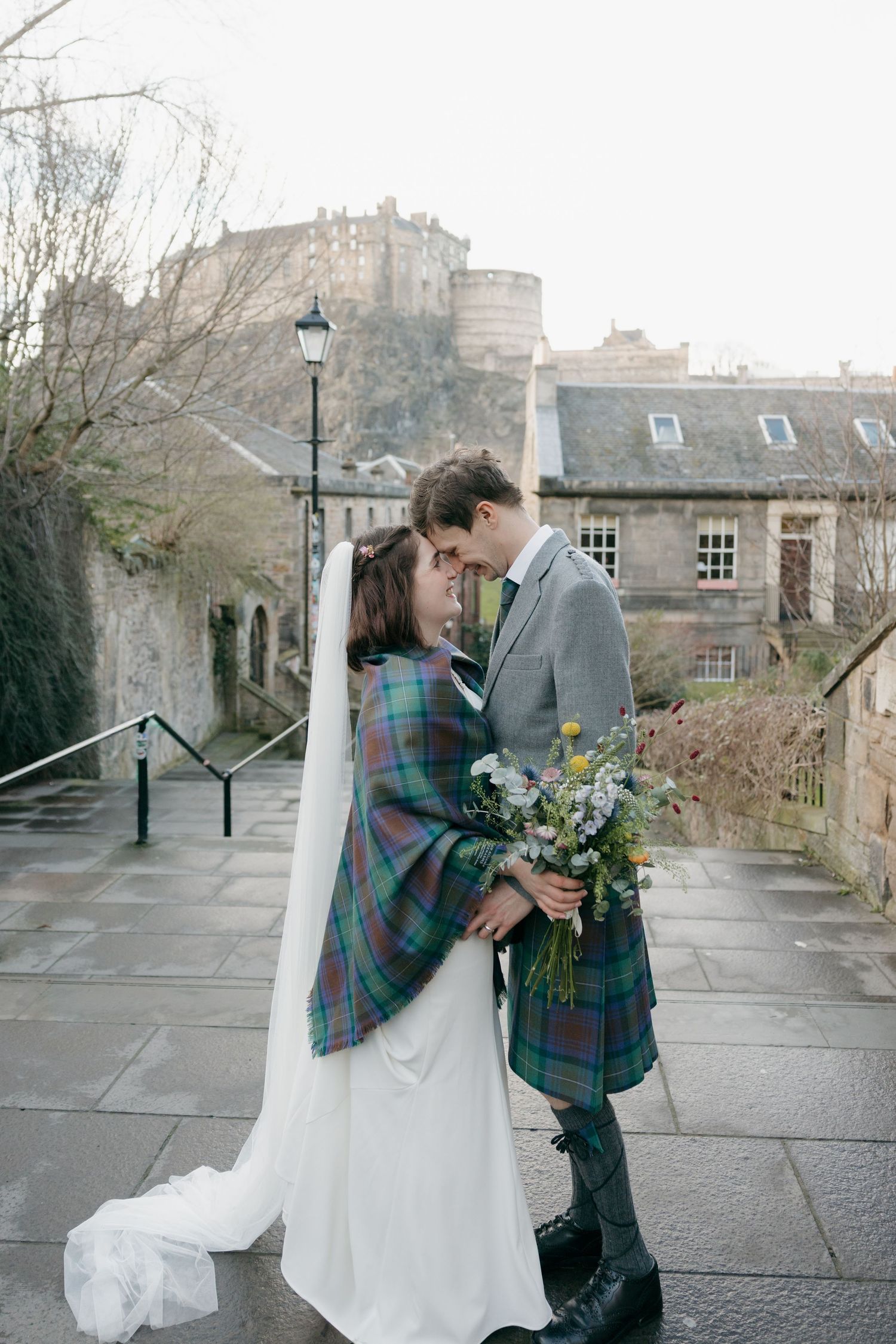 Bride and groom in Scottish tartan embrace on cobblestone steps with Edinburgh Castle in the background.