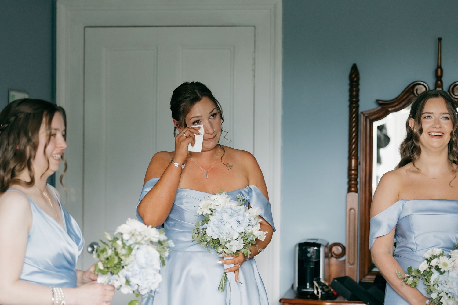 Three bridesmaids in light blue off-shoulder gowns holding white bouquets, one wiping tears of joy.