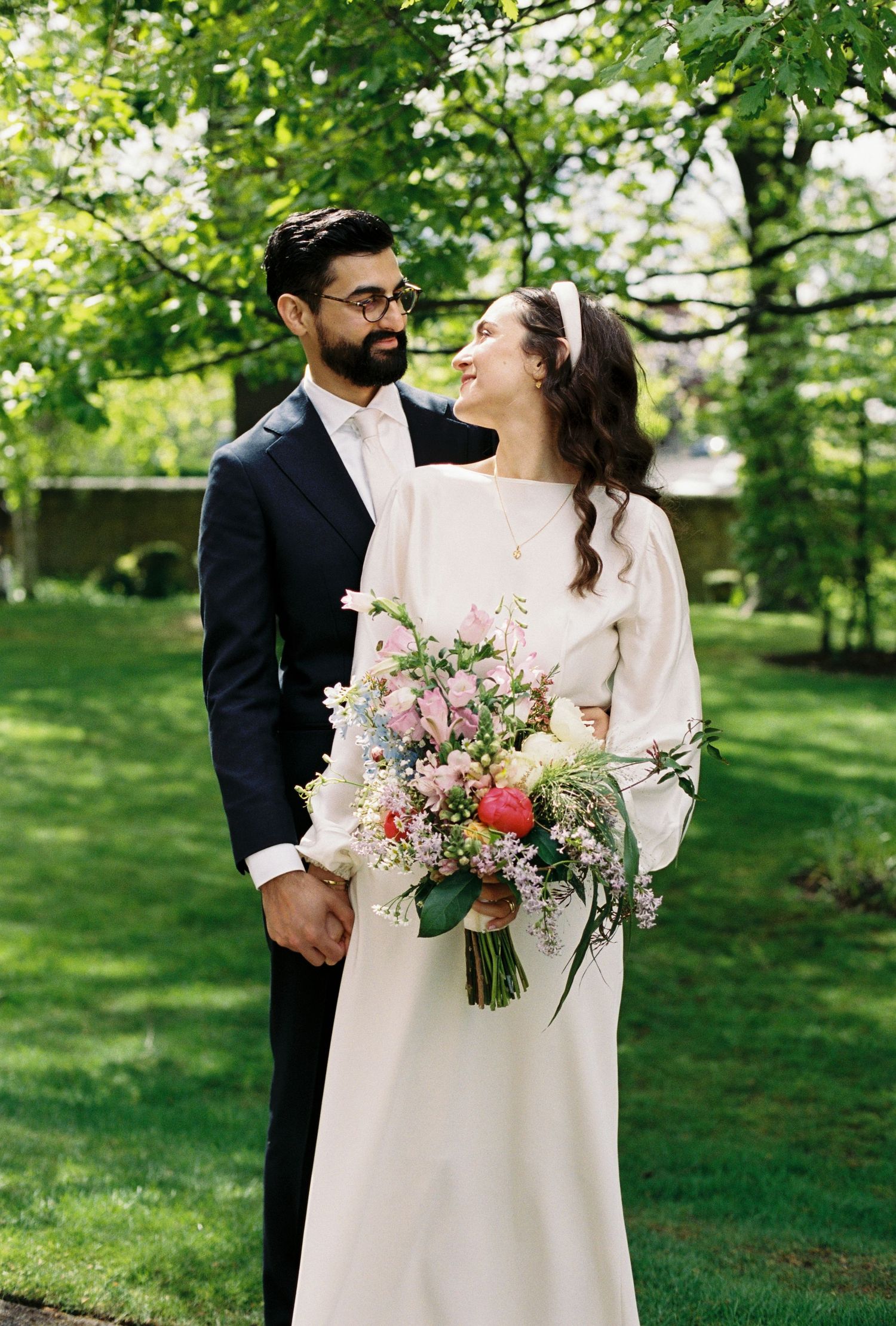 Couple in wedding attire embracing outdoors, groom in dark suit, bride in white gown holding colorful spring bouquet.
