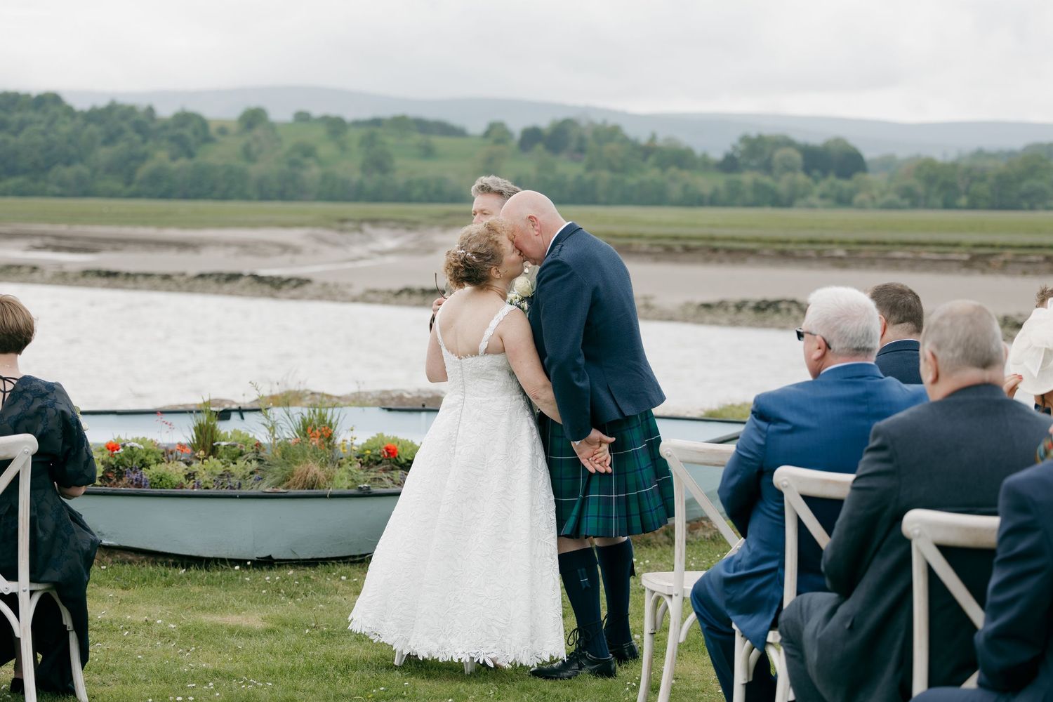 Bride and groom share first kiss at outdoor Scottish wedding ceremony by a loch, groom wearing traditional kilt.