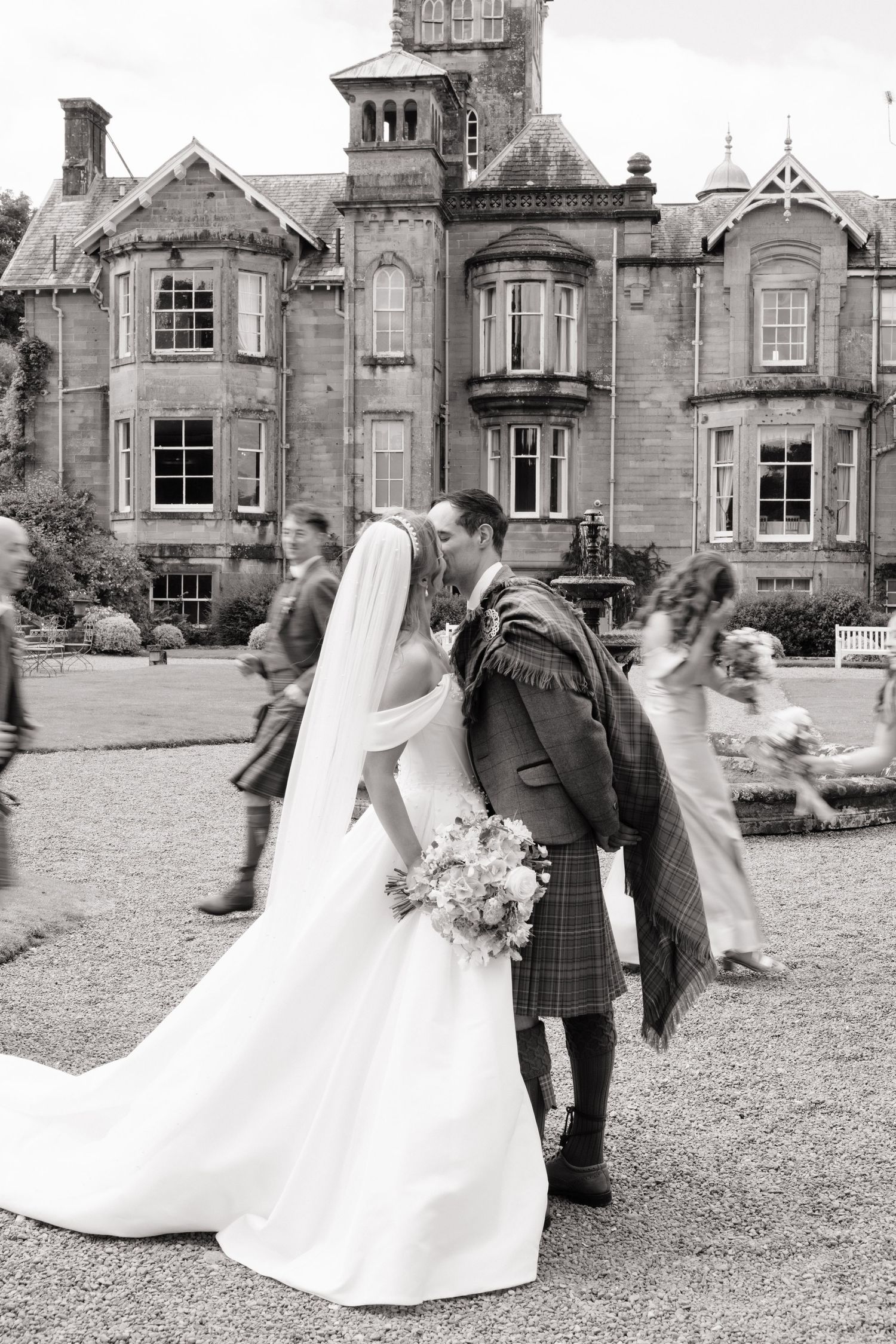 Black and white photo of bride and groom kissing in kilts outside a grand Scottish manor house wedding venue.