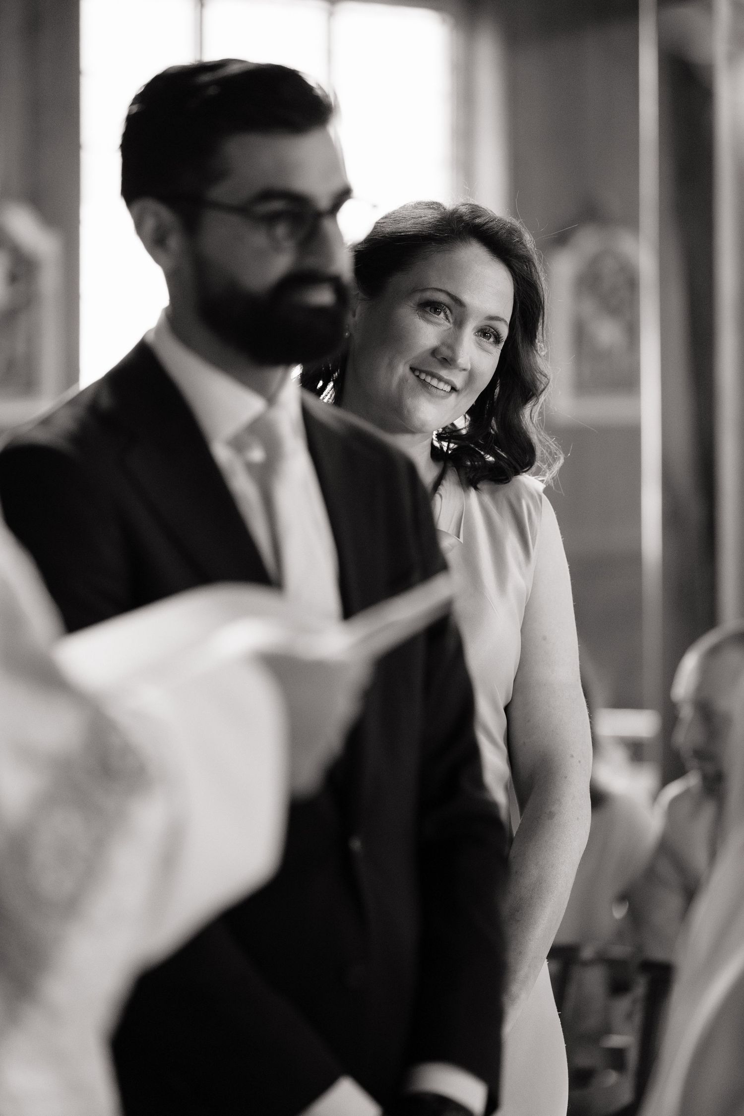 Black and white photo of a smiling bride and bearded groom in formal attire during a wedding ceremony.