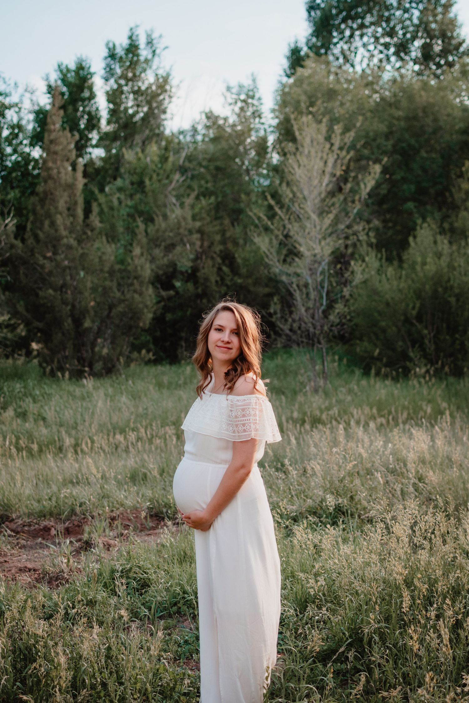A person in a flowing white dress stands in a sunlit meadow surrounded by tall grass and trees.