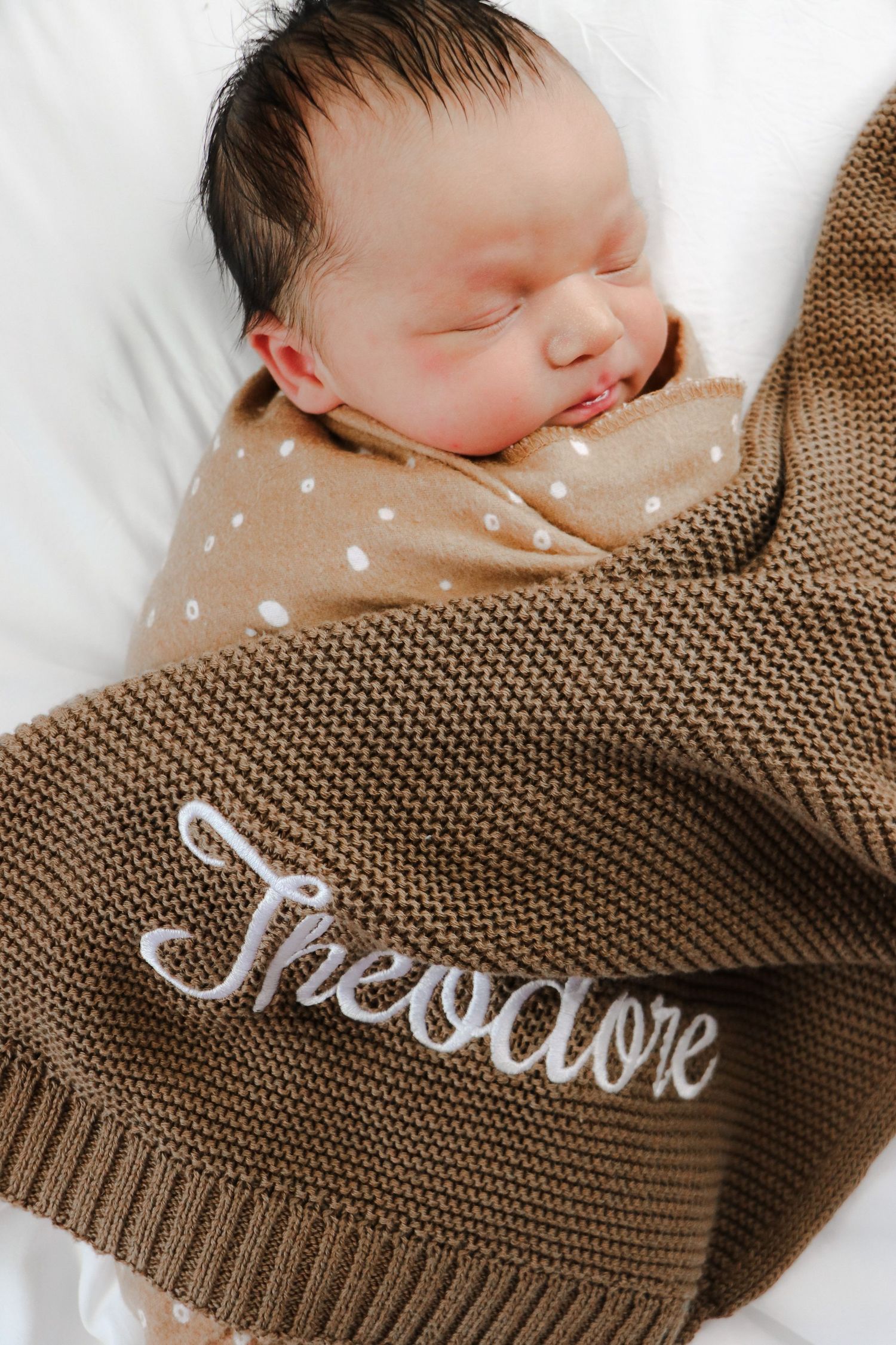 Brown knit blanket with embroidered name being used by sleeping baby in white bedding.