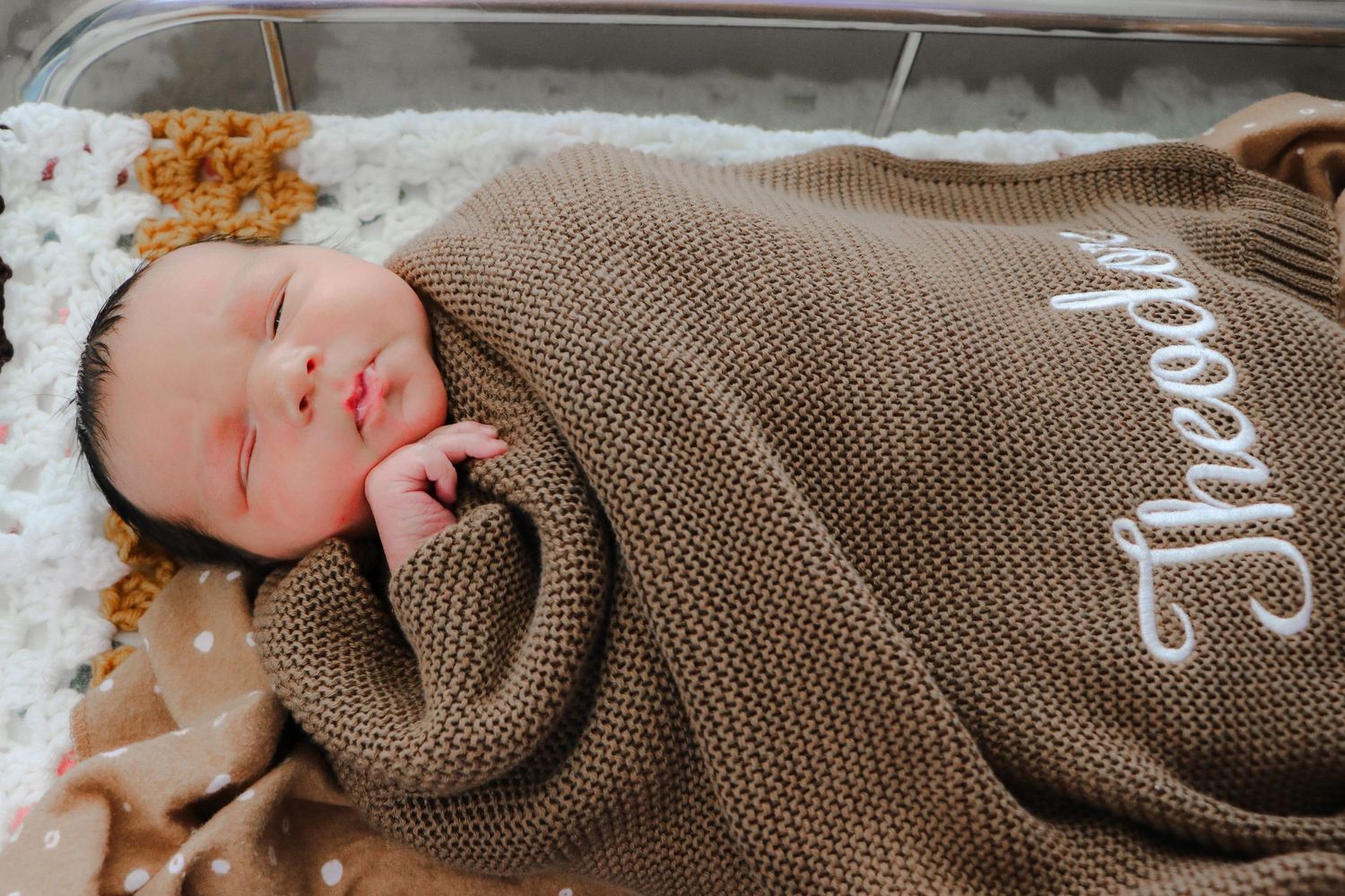 A baby wearing a cozy brown knit sweater lies peacefully on a white surface.