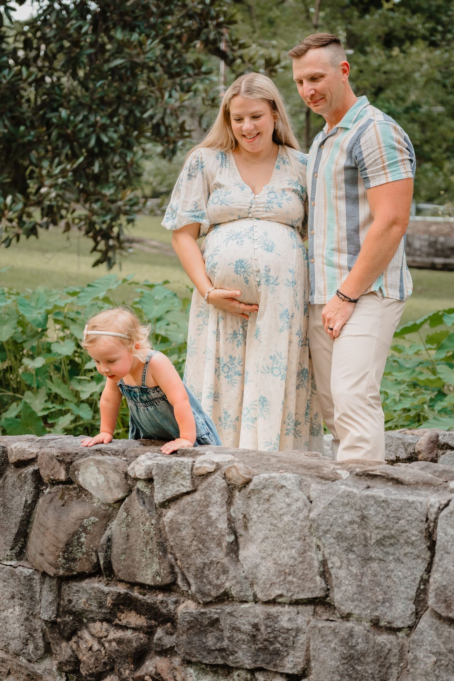 Pregnant woman in floral dress with husband and toddler daughter posing on stone wall outdoors.