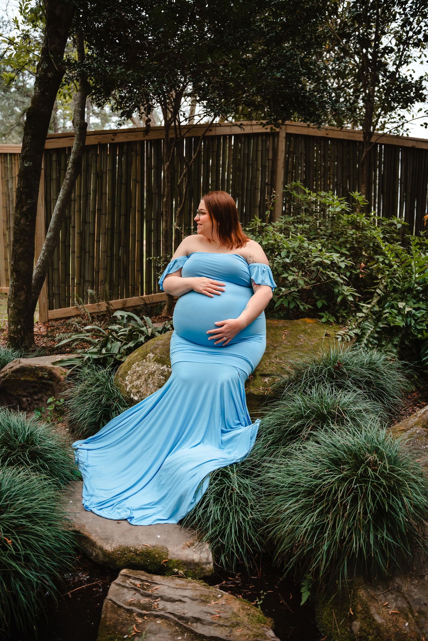 A pregnant woman in a blue dress stands in a garden surrounded by greenery and a wooden fence.
