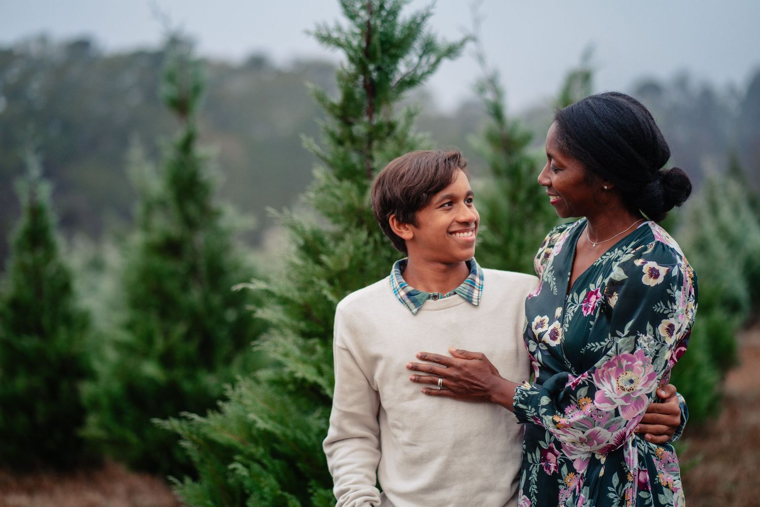 A couple shares a tender moment while standing among evergreen trees in a natural outdoor setting.