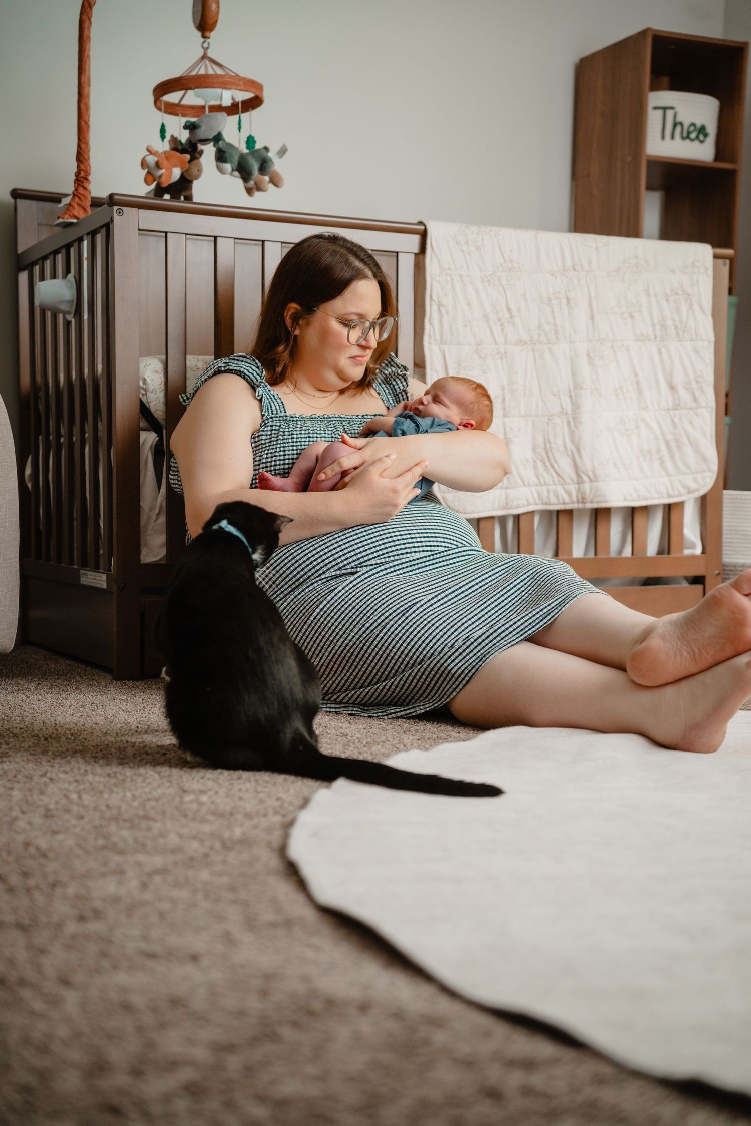 Mother sitting on nursery floor holding newborn baby while black Labrador dog looks on beside wooden crib.