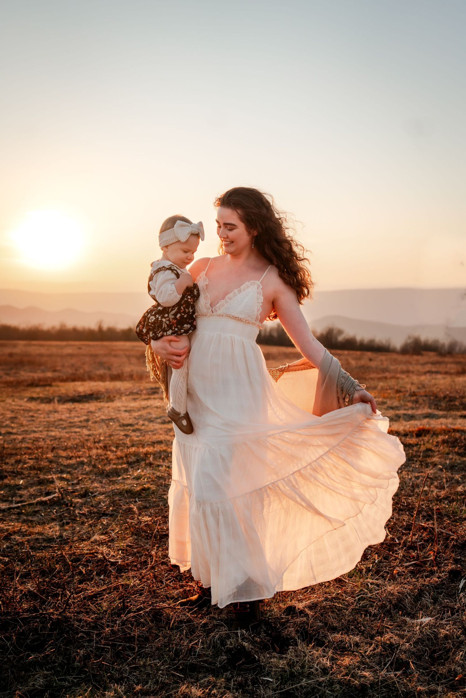 A mother in a white dress holds her baby at golden sunset in an open field, her skirt flowing in the breeze.