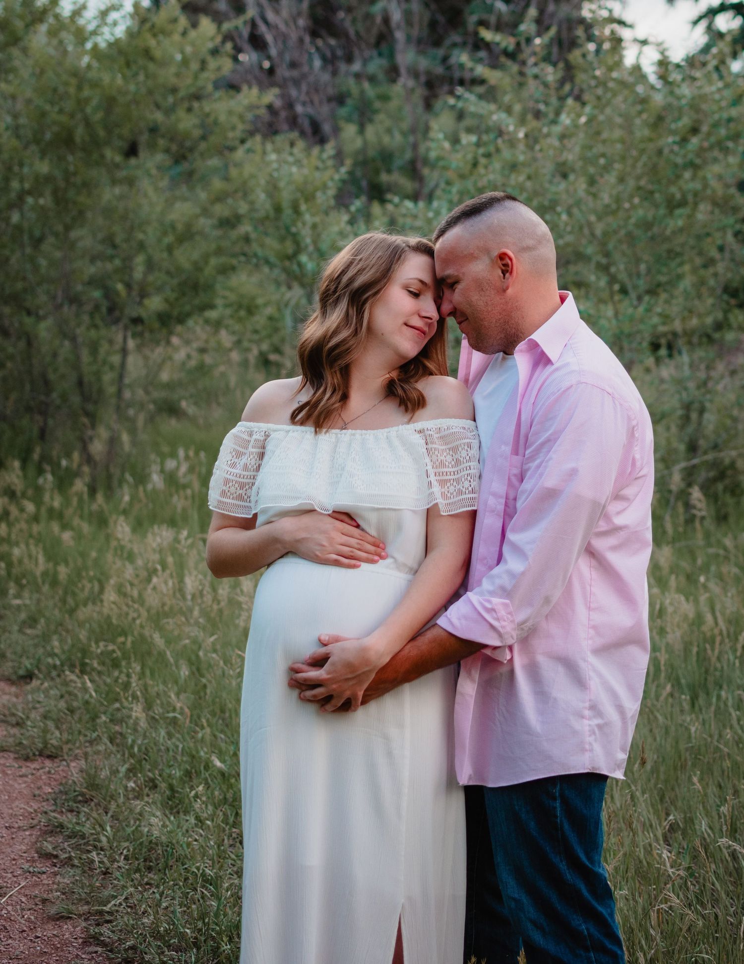 A tender maternity photoshoot in a natural outdoor setting with a couple wearing white and pink.