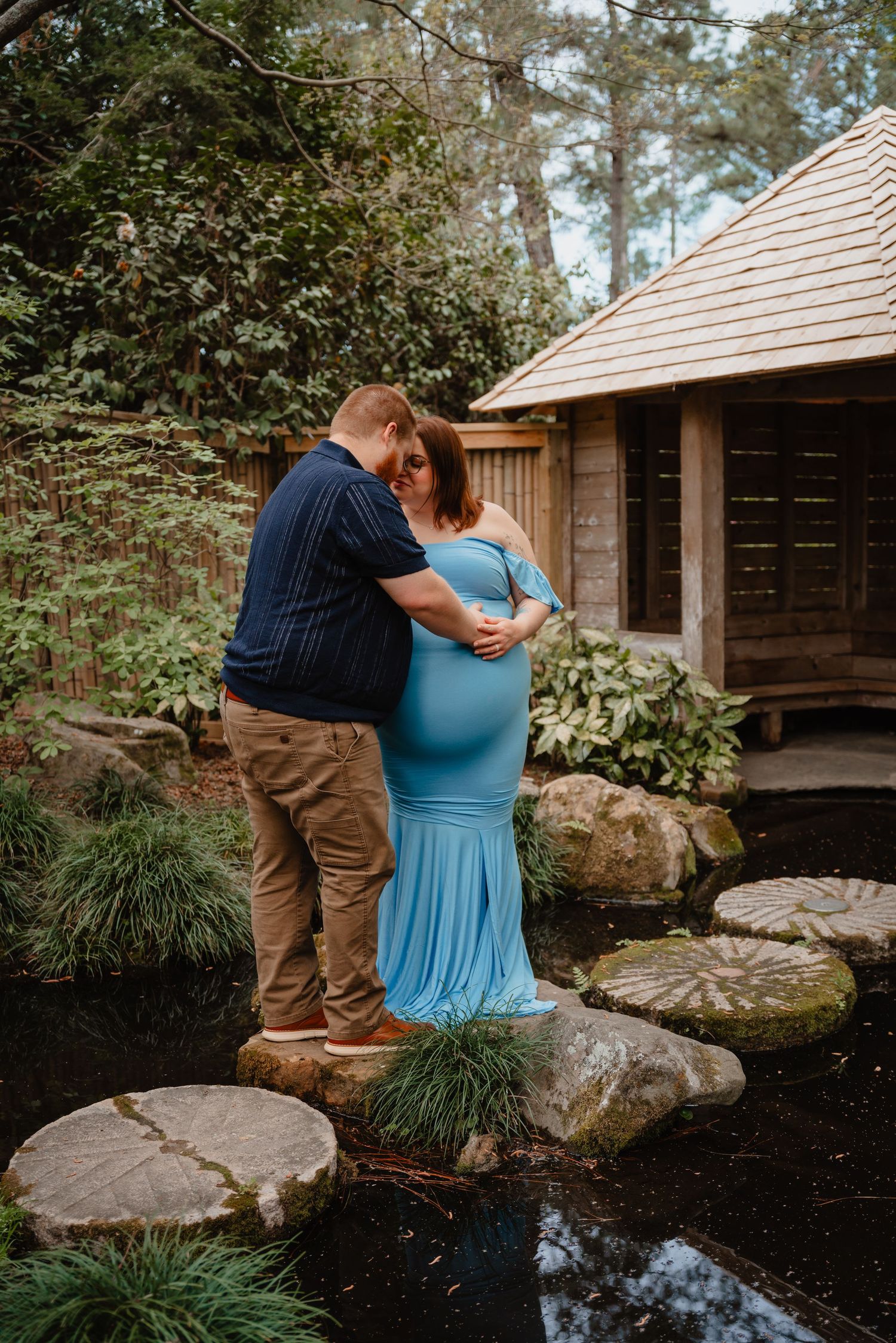 A couple embraces in a rustic wooden cabin surrounded by lush greenery and a small stream.