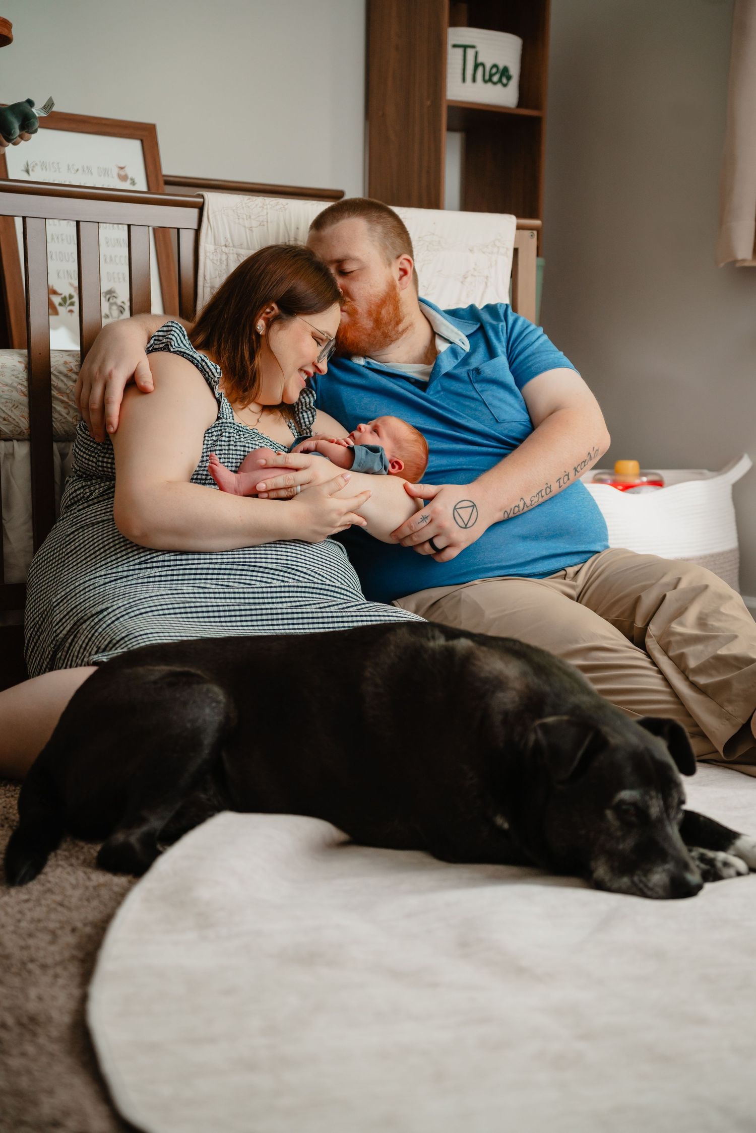 New parents sit on nursery floor holding newborn baby while black Labrador rests nearby, crib with name Theo visible behind them.