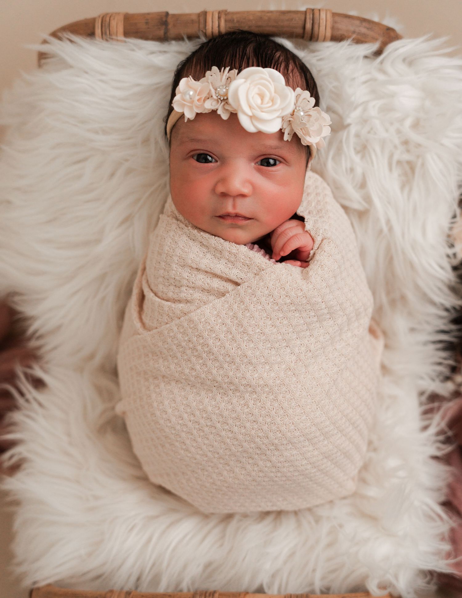 Newborn baby girl swaddled in cream knit wrap, wearing white floral headband, lying on fluffy white fur blanket.
