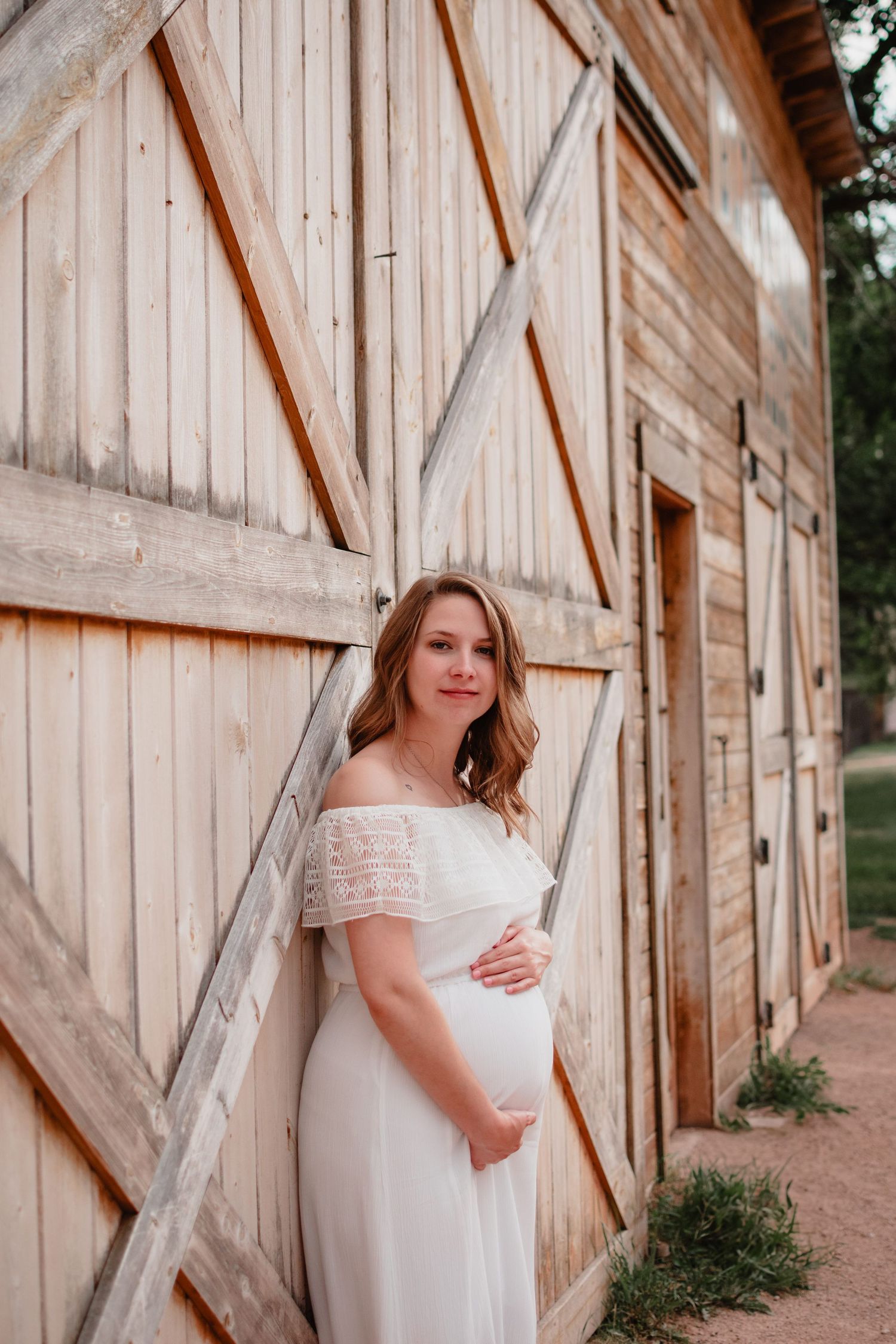 Rustic maternity portraits against a weathered wooden barn wall showcasing an elegant white lace dress.