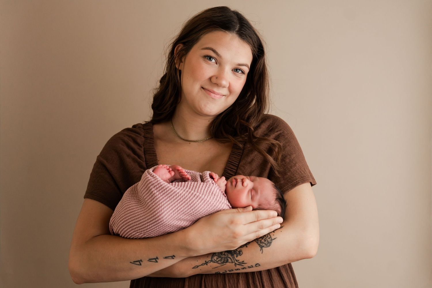 A young mother with tattoos holds her sleeping newborn wrapped in pink in a studio portrait.