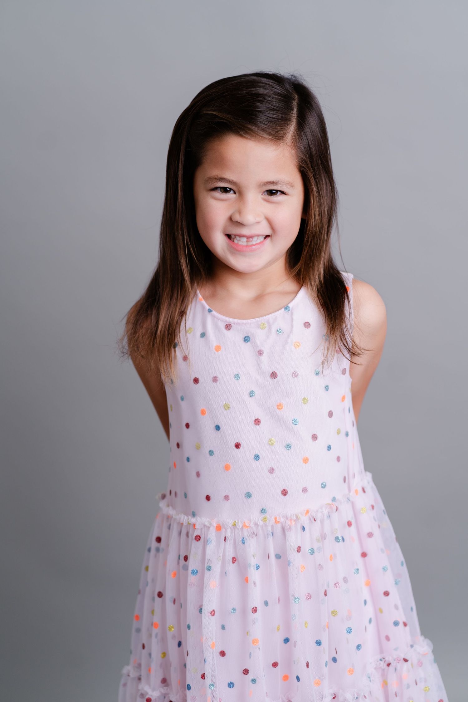 Child wearing a white polka dot dress poses against a gray background in a series of playful expressions.