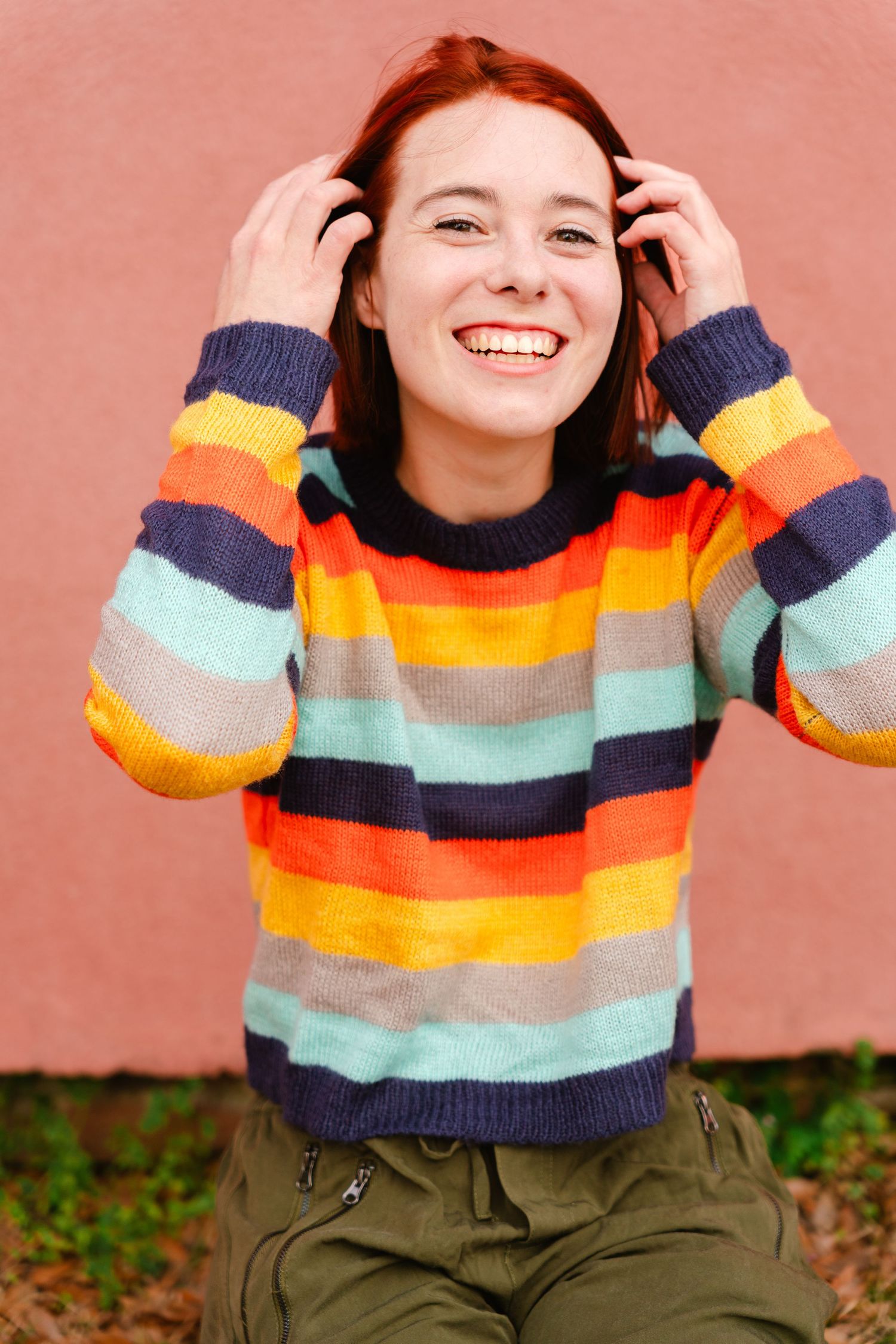 Playful poses in striped rainbow sweater against coral colored wall.