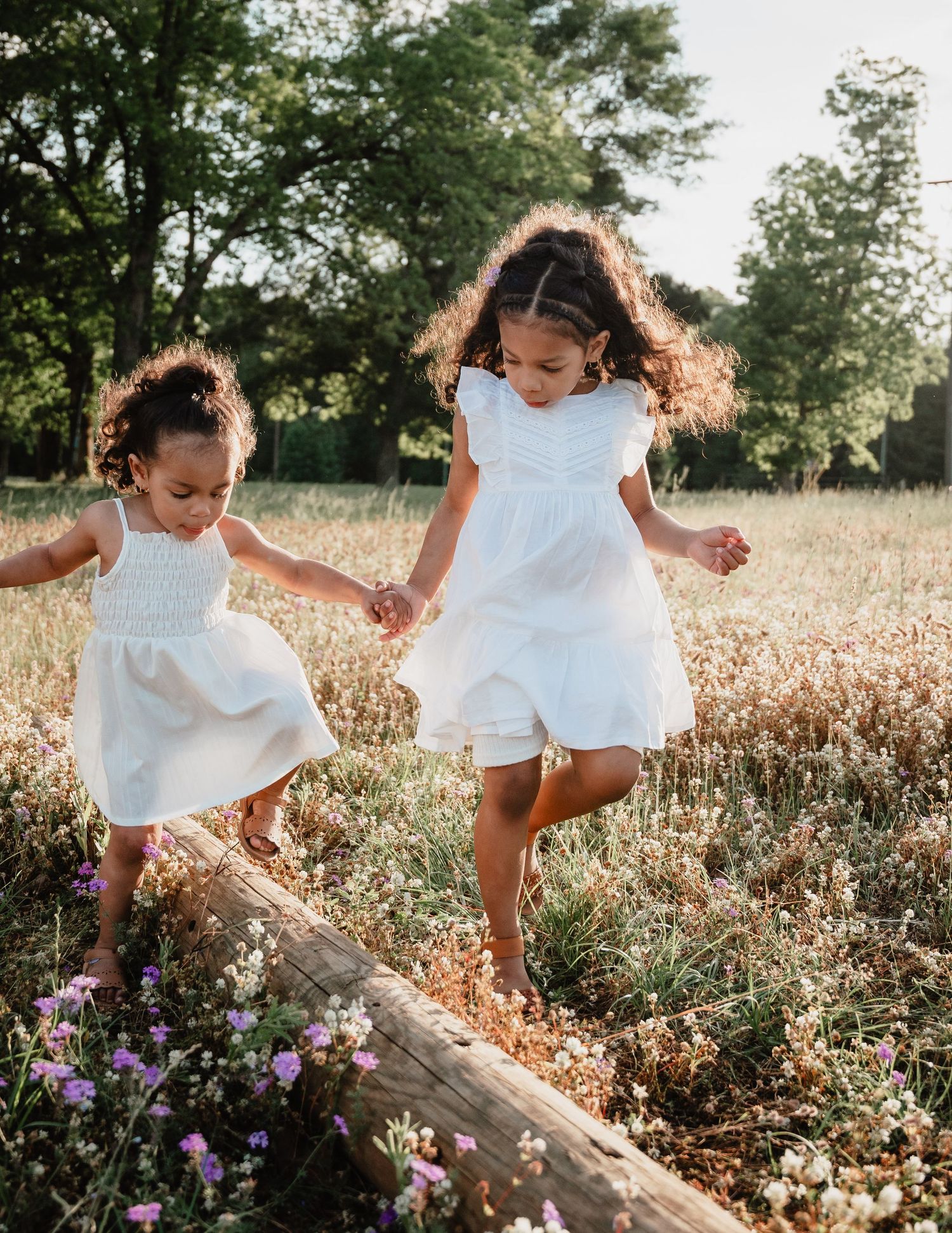 Two people in white dresses dance joyfully through a sunlit field of flowers.